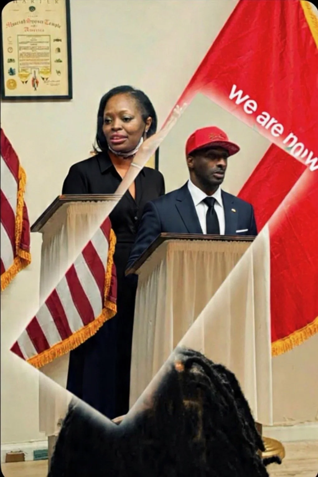 Two individuals standing at podiums during a ceremony, with American flags and a red flag that reads 'we are now' partially visible. The woman is wearing a black outfit and a face mask around her neck, and the man is wearing a navy suit, white shirt, black tie, and a red cap.