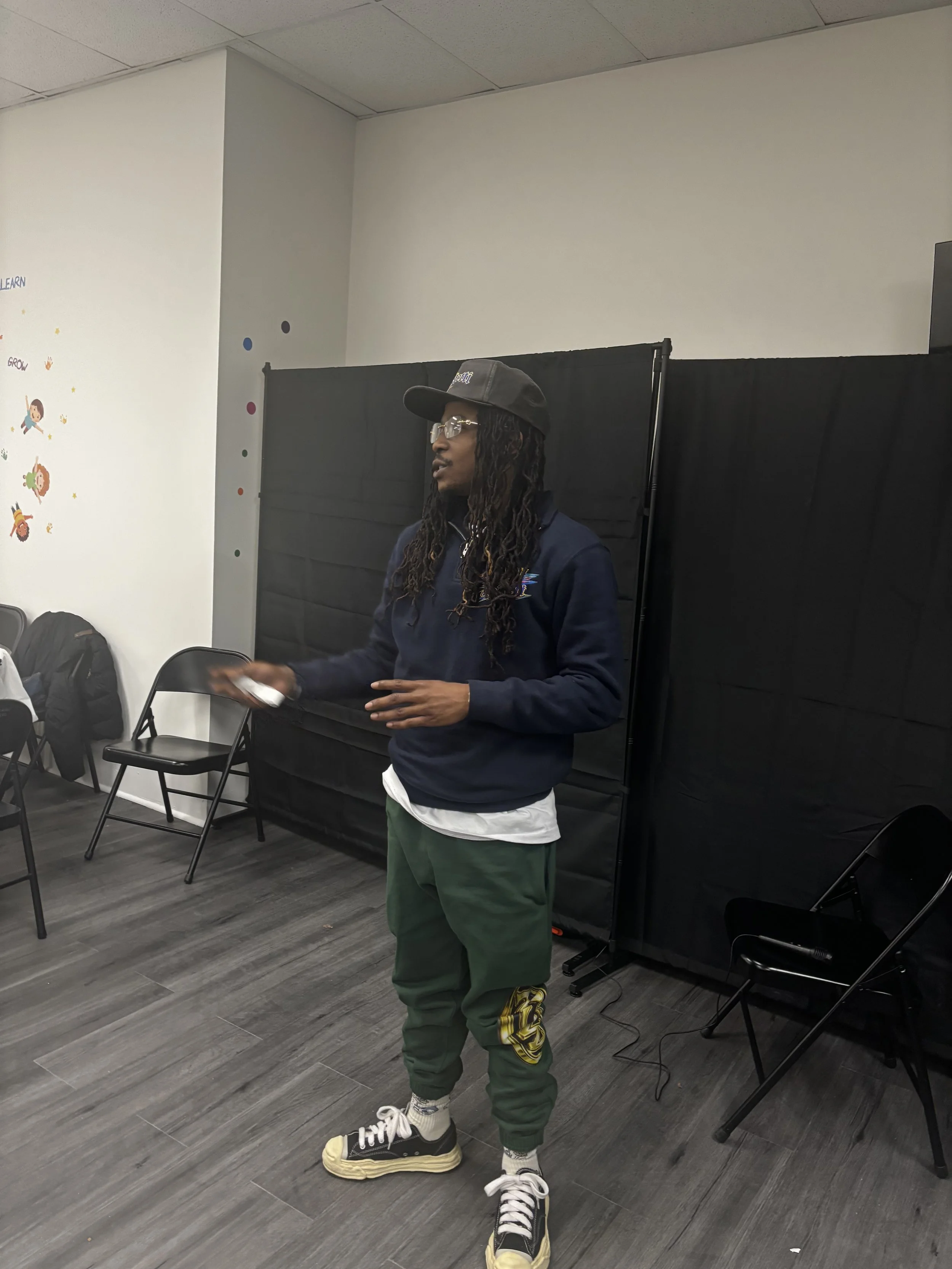 A young man with dreadlocks, glasses, a black cap, navy hoodie, green pants with a yellow logo on the thigh, and sneakers standing in a room with black partitions, chairs, and colorful wall decorations.
