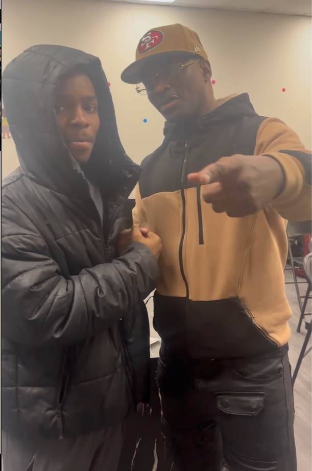 Two individuals posing indoors, one wearing a black puffer jacket with a hood, the other wearing a black and tan zip-up hoodie, a yellow baseball cap, and glasses, pointing at the camera.