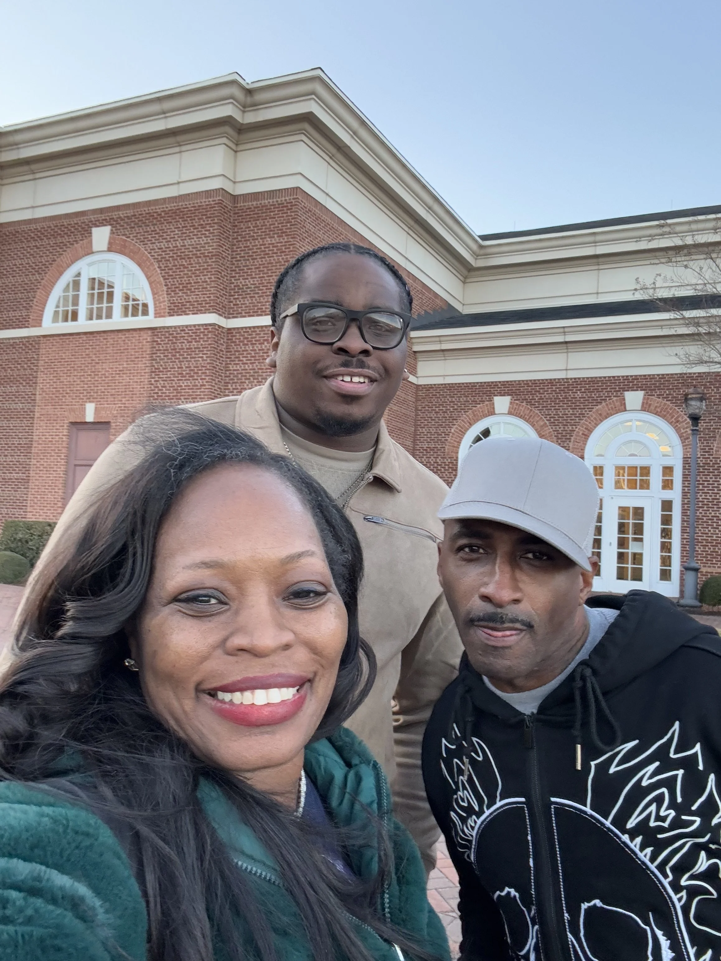 A group of four people taking a selfie outside in front of a brick building with arched windows, under a clear blue sky.