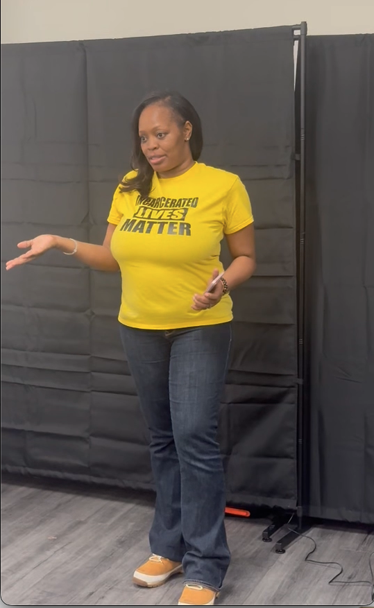 A woman standing and gesturing with her right hand in front of a black backdrop. She is wearing a yellow T-shirt with black text, blue jeans, and tan shoes. She holds a phone in her left hand.