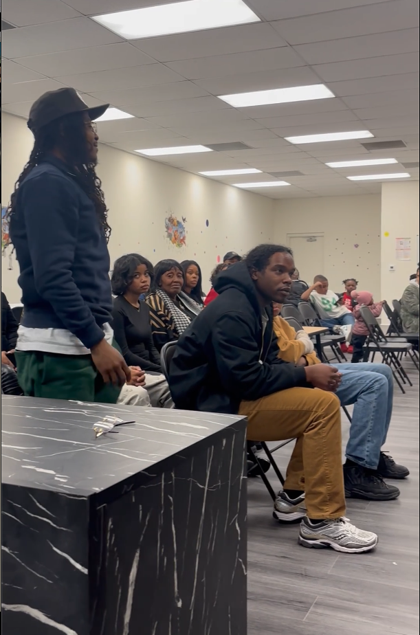 A group of young people seated and one standing in a room with plain walls and ceiling lights, appearing attentive or engaged in an event.