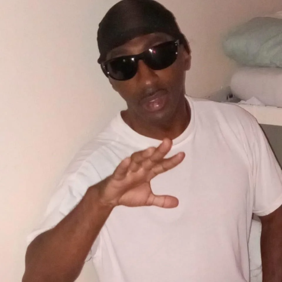 Person wearing black sunglasses and a white t-shirt, making a hand gesture indoors.