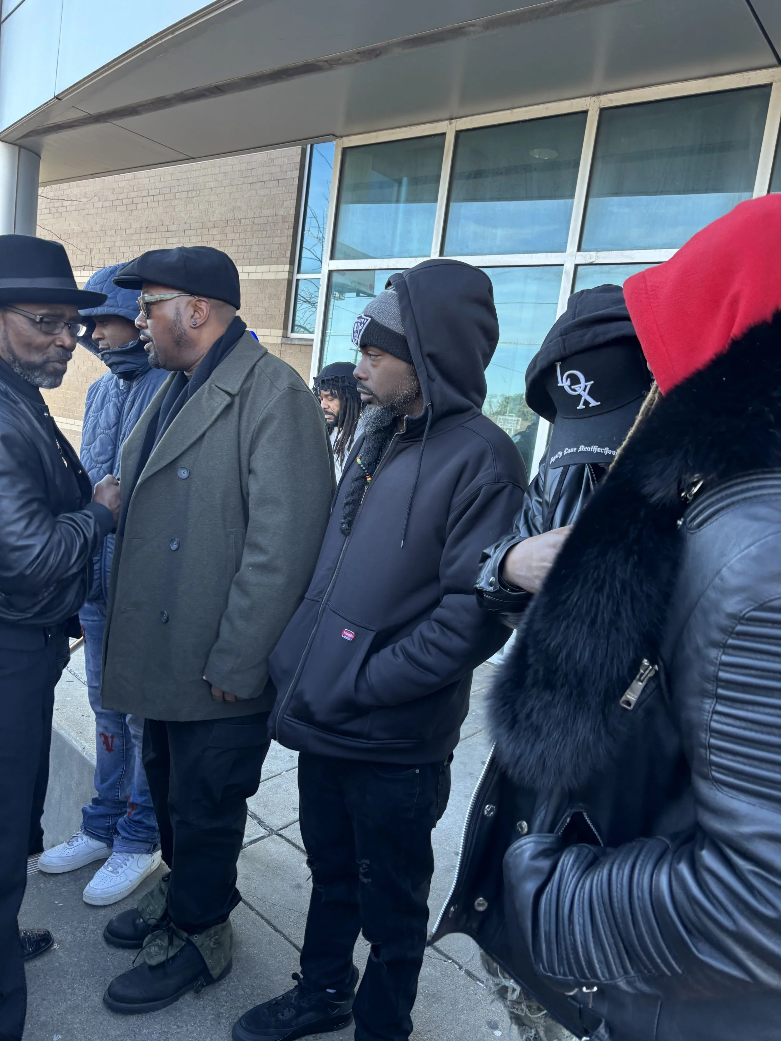 A group of five men standing outdoors, engaging in conversation, wearing winter clothing such as jackets, hoodies, and hats, with two wearing glasses, and one with dreadlocks, in front of a building with large windows.