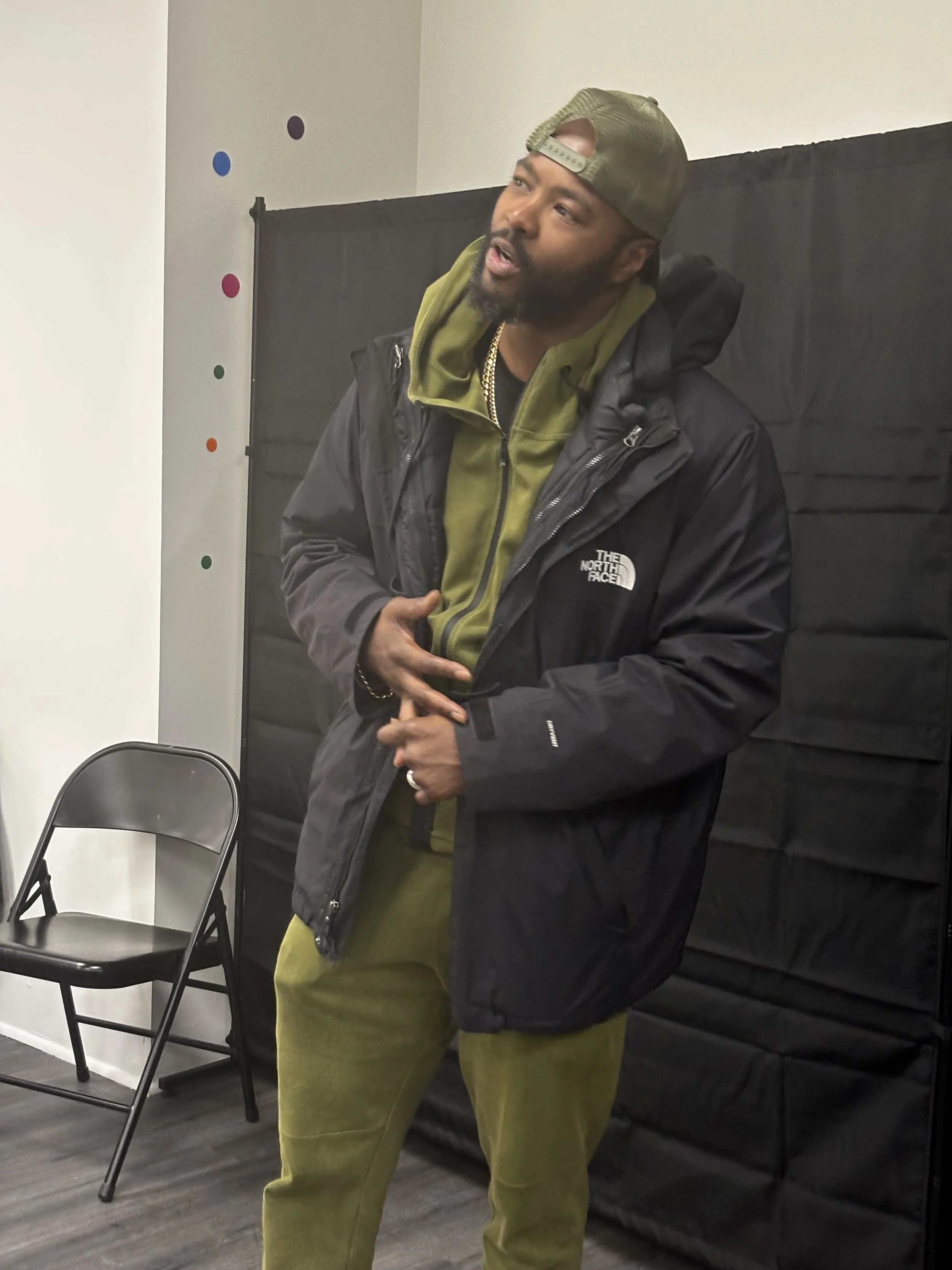 A man with a beard wearing a green baseball cap backwards, a black North Face jacket, a gold chain, and green pants, standing indoors against a black backdrop, looking slightly upward and appearing to speak or sing.