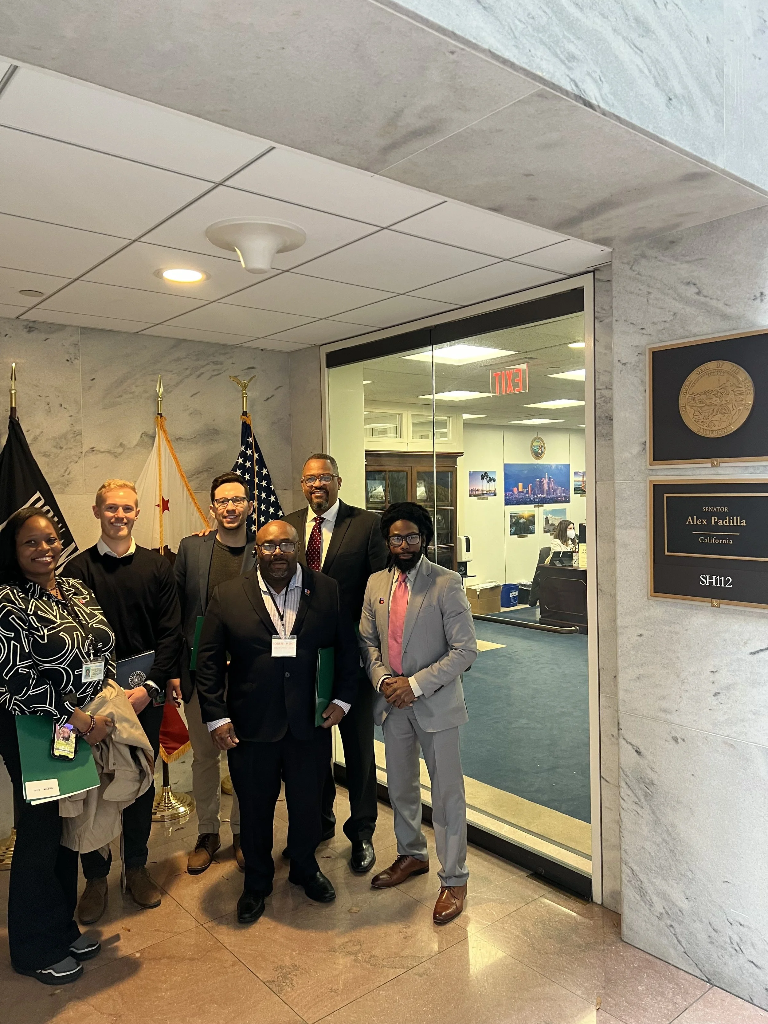 Group of people standing in front of a government building with flags and a plaque honoring a California senator, with office interior visible through glass doors.