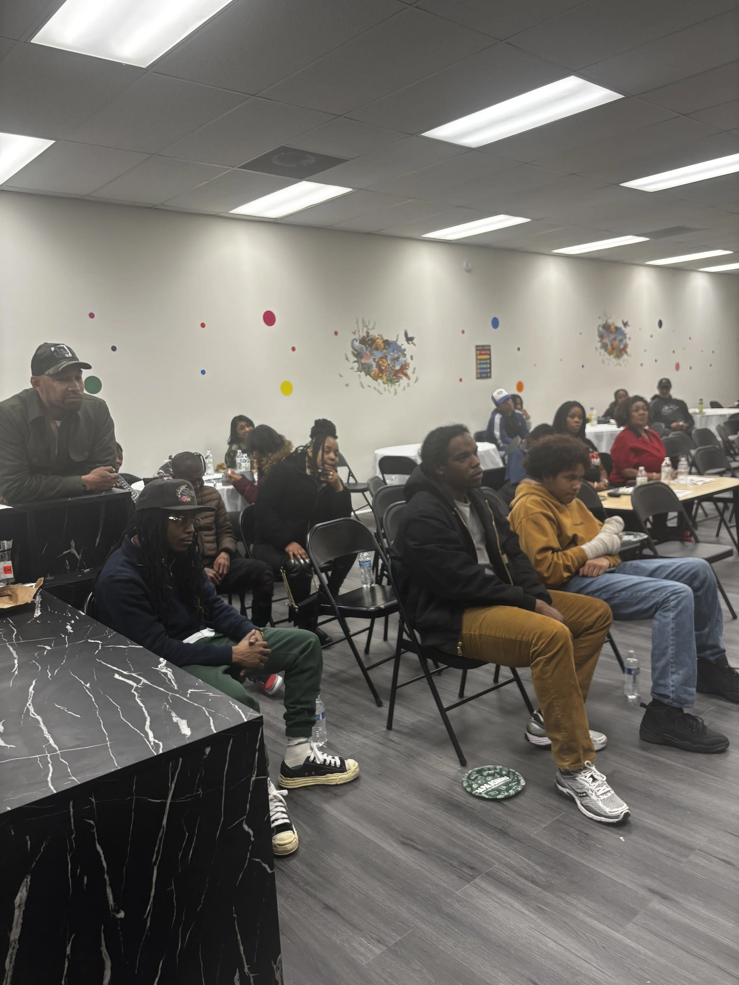 A group of people sitting in a conference room during a meeting or event, with some looking attentively towards the front. The room has white walls with colorful dot decorations and a mural on the wall, with tables, chairs, and bottled water.
