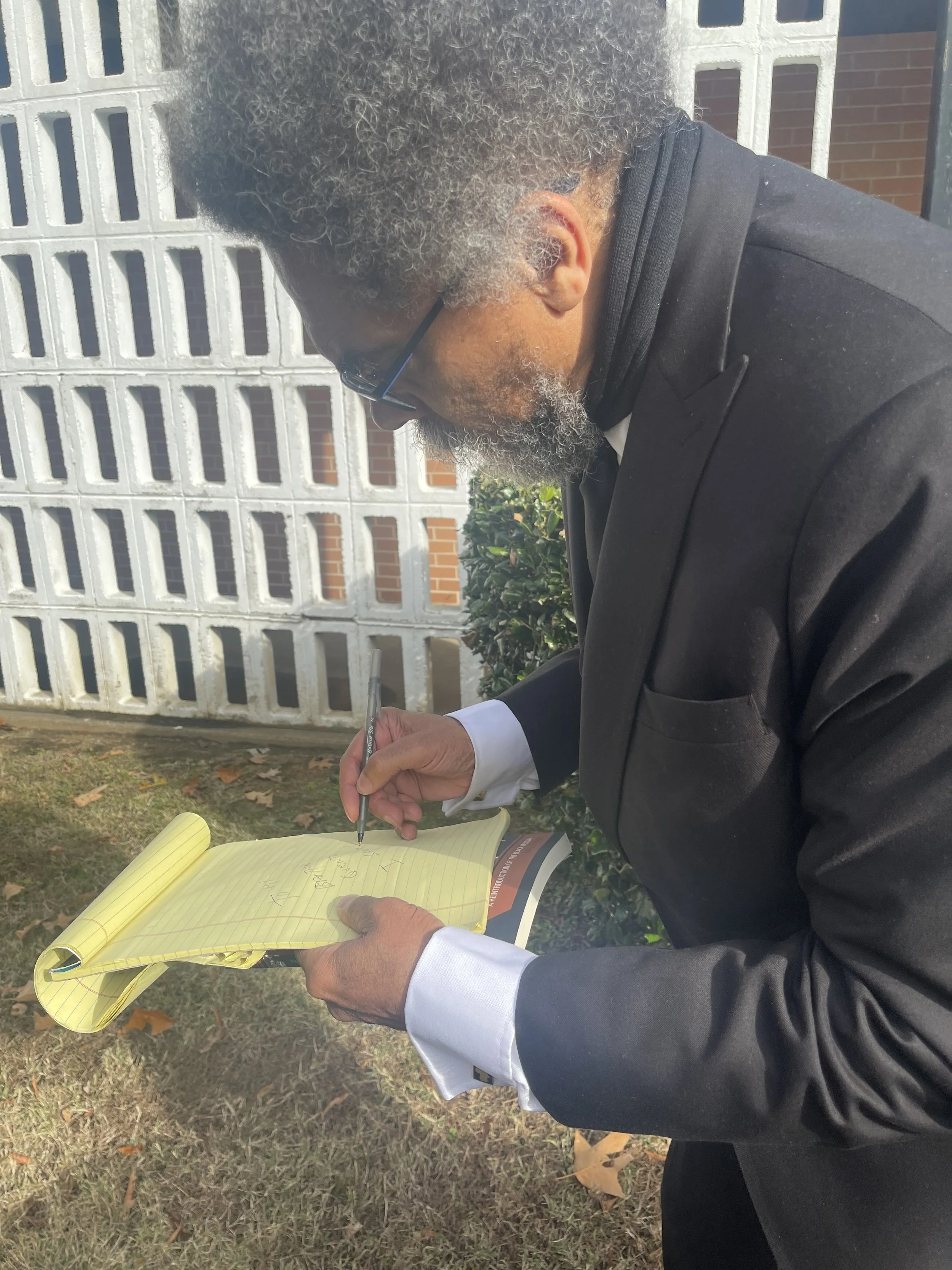 An older man with gray curly hair, wearing glasses and a black suit, is writing in a yellow notepad outdoors, near a fence and brick building.