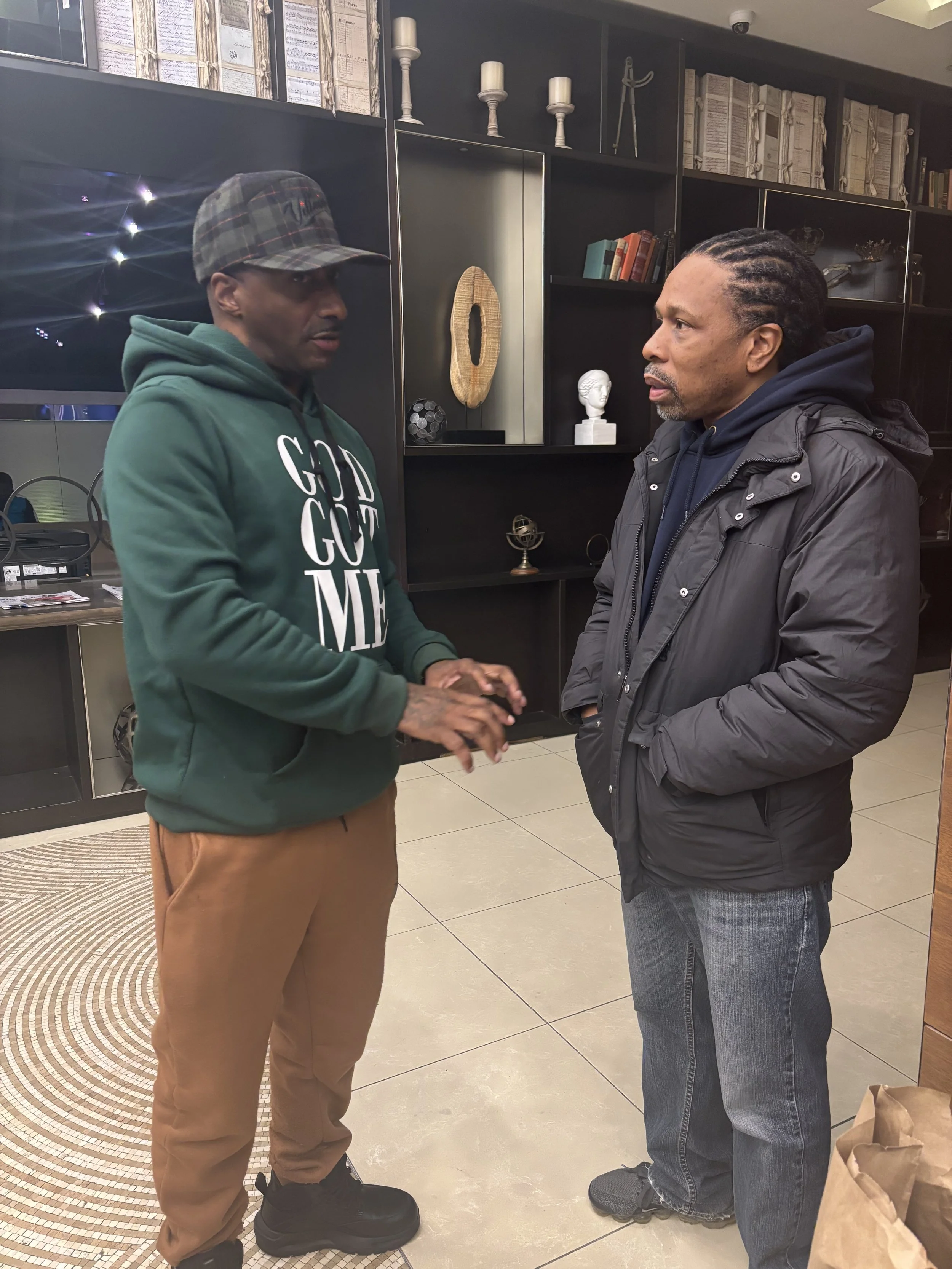 Two men are standing face to face indoors, engaging in conversation. The man on the left wears a green hoodie with white lettering, tan pants, and a plaid cap. The man on the right wears a dark jacket and jeans, with braided hair. Behind them, there is a black shelving unit with decorative items, books, and a bust sculpture.