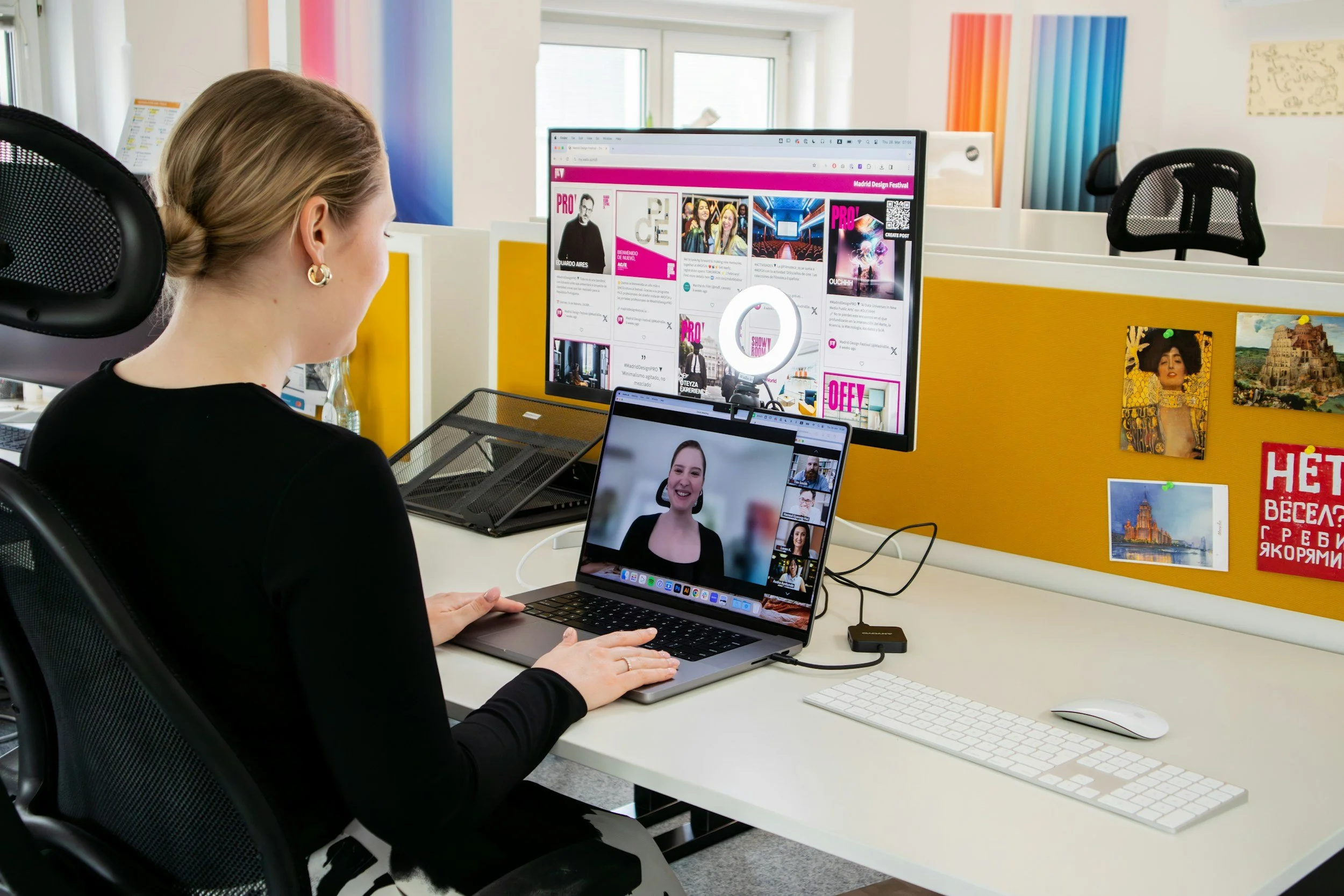 A woman with blonde hair wearing earrings, sitting at a desk in an office, participating in a video call on her laptop and desktop monitor. The office includes a colorful division with pictures and posters on a yellow and white partition, and a black office chair with a mesh backrest.