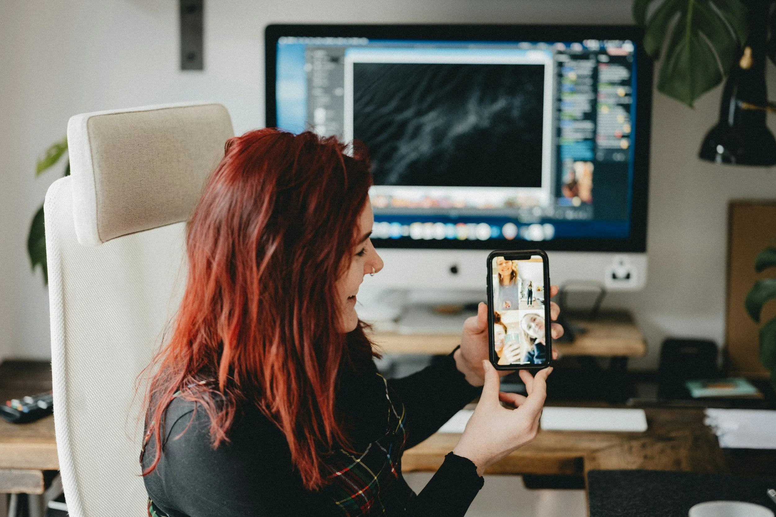 A woman with red hair sitting at a desk taking a selfie with her smartphone, which shows her face on the screen. She is in front of a computer monitor and an office environment.