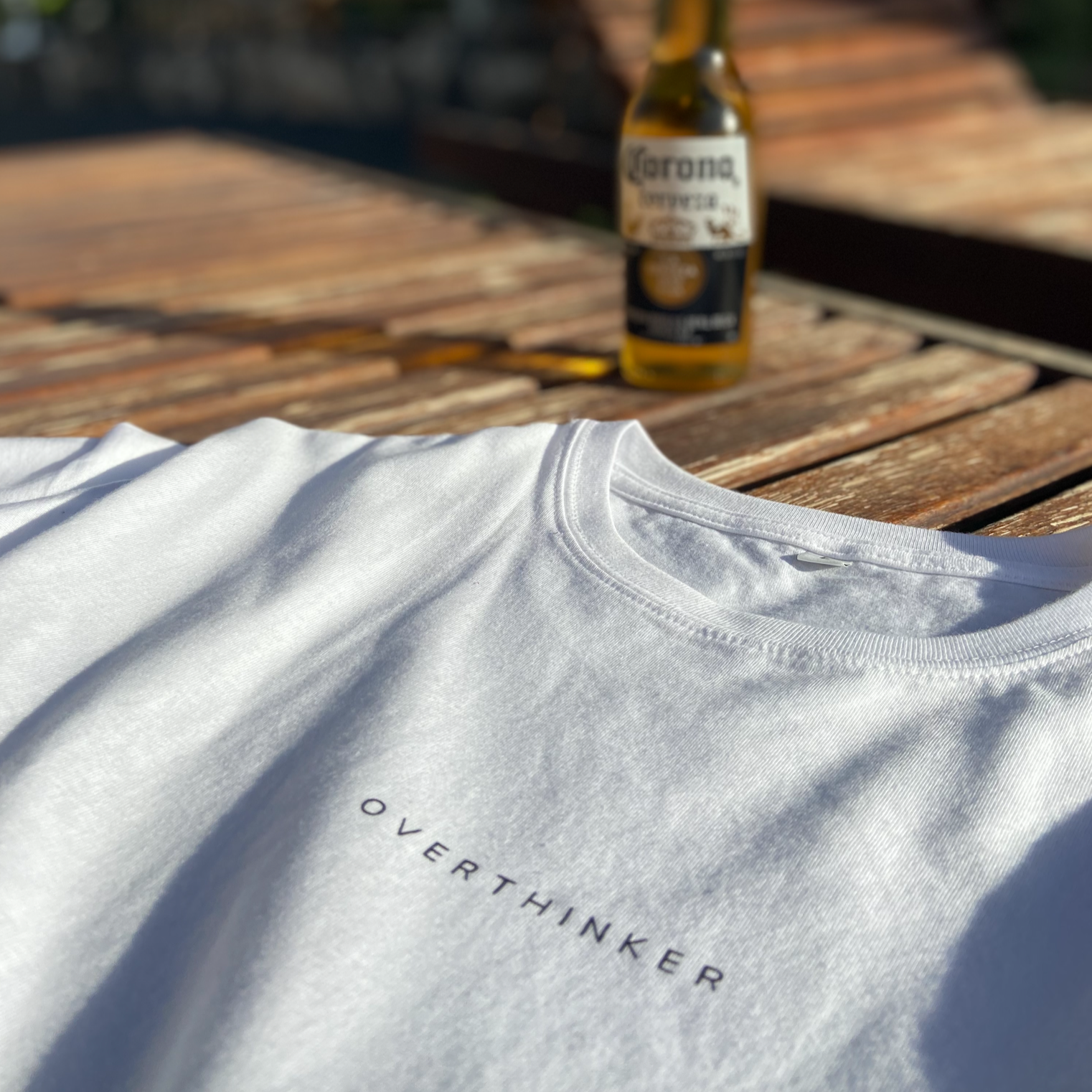 A white T-shirt with the words "OVERTHINKER" printed on it, laid on a wooden surface outdoors. In the background, there is a blurred bottle of Corona beer.