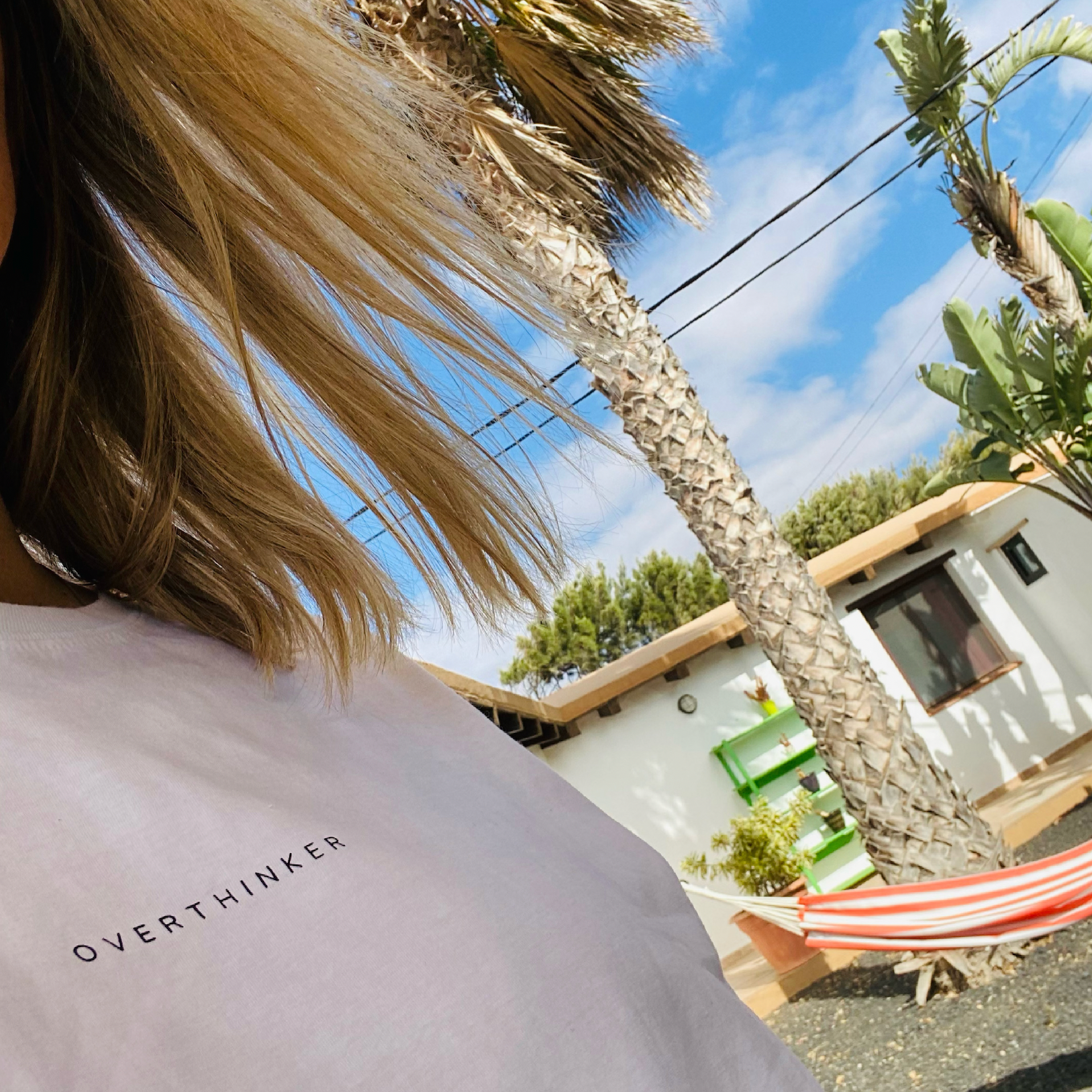 Partial view of a person wearing a white shirt with black text, standing outdoors near a white house with a brown roof, palm trees, green plants, and a hammock, against a background of blue sky and some clouds.
