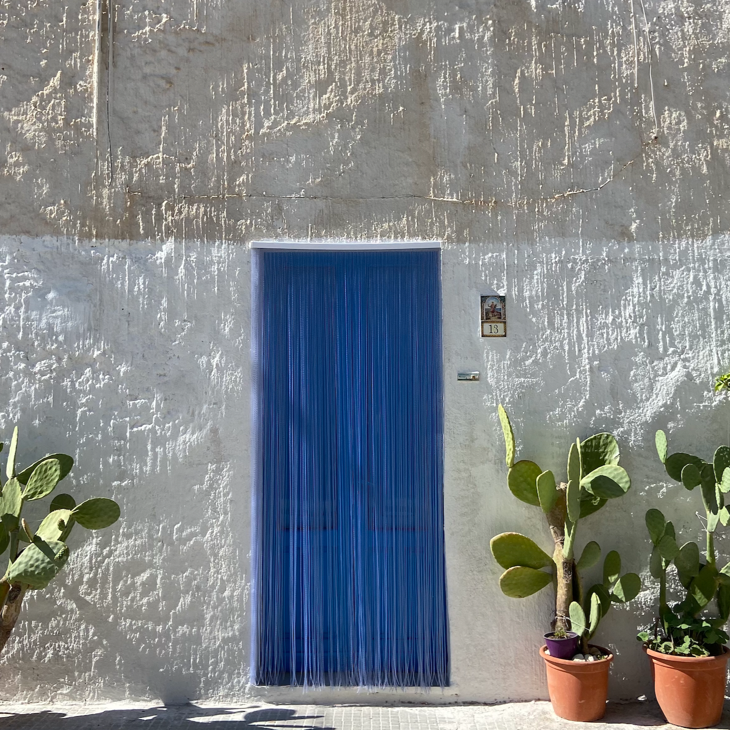 White textured stucco wall with a blue door and house number 13. Two potted cactus plants are in front of the wall, and there is a small religious picture above the house number.