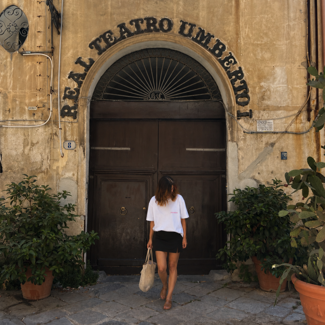 A woman with brown hair, wearing a white t-shirt and black mini skirt, carries a woven bag, walking out of an old building with a large wooden door and plants in pots outside. The building has a sign that reads 'Teatro Umberto' above the entrance.