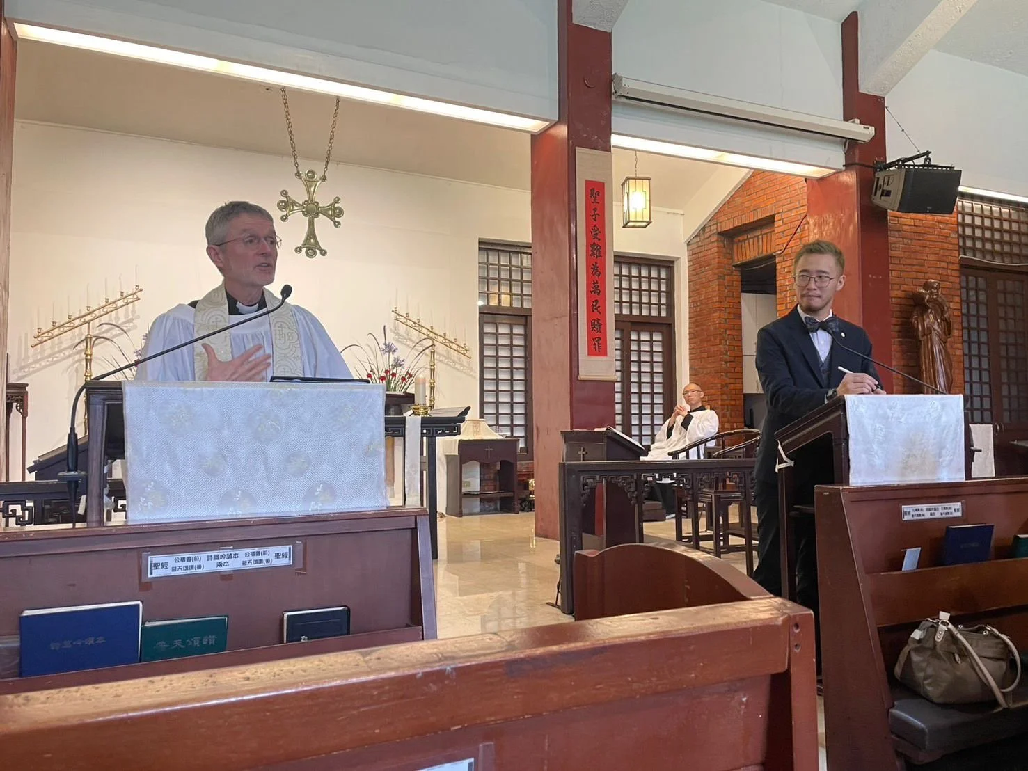 A priest and a young man in tuxedo giving a speech at a church service, with a seated clergyman in the background, inside a church with wooden pews, religious statues, and Asian-style decor.