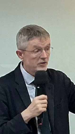 A man with gray hair and glasses speaking into a microphone, wearing a dark suit and light blue shirt.