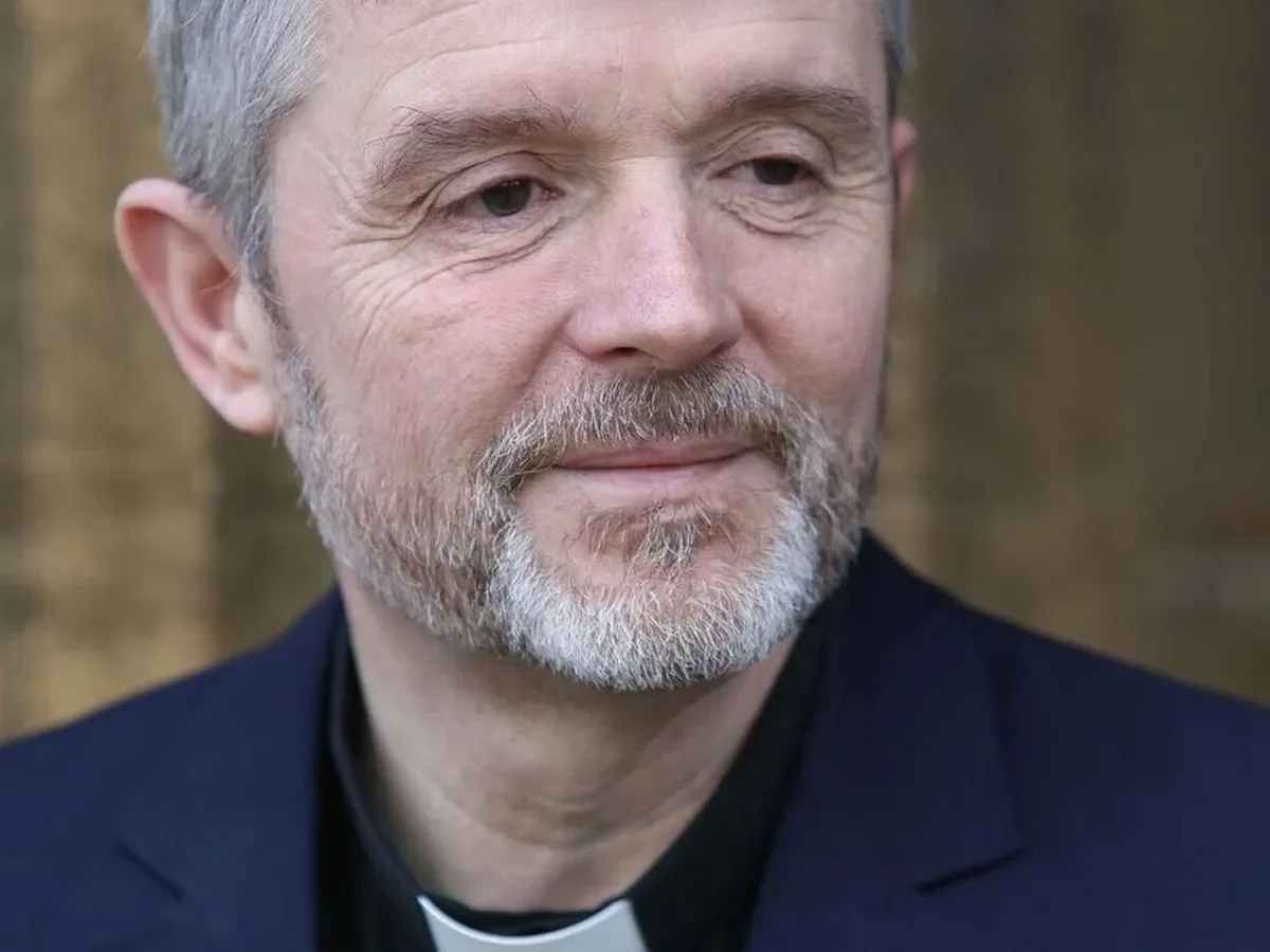 Close-up of a middle-aged man with a beard and mustache, wearing clerical attire and a black shirt underneath, standing outdoors with a blurred background.