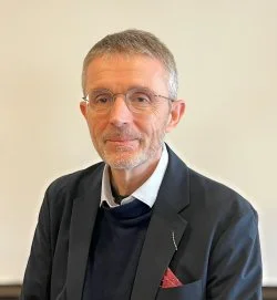 A middle-aged man with glasses, short gray hair, and a light beard, wearing a dark blazer over a navy sweater, standing in front of a plain light-colored wall.