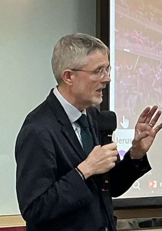 A man with gray hair and glasses speaking into a microphone in front of a large screen, wearing a dark suit and tie.