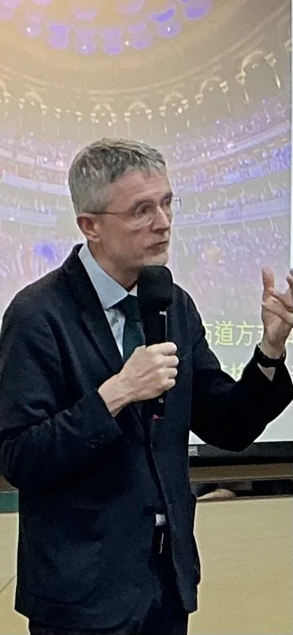 A man in a dark suit and glasses speaking into a microphone in front of a projected screen with a large audience in a theater or conference hall.