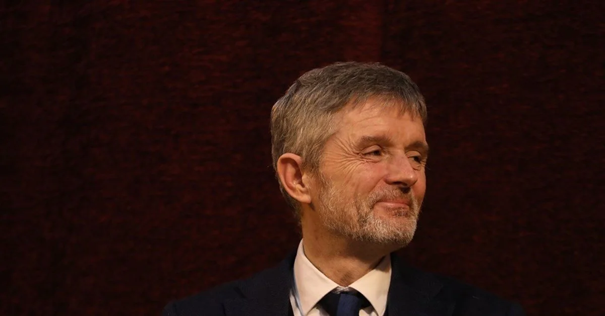 A middle-aged man with gray hair and beard, wearing a dark suit and tie, smiling and looking to his right, against a dark red background.