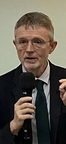 A man in a suit and glasses speaking into a microphone during a speech or presentation