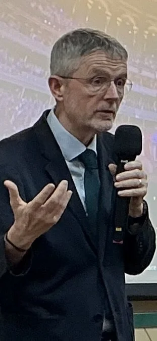 A middle-aged man with gray hair and glasses speaking into a microphone, wearing a dark suit and tie, during a presentation or speech.