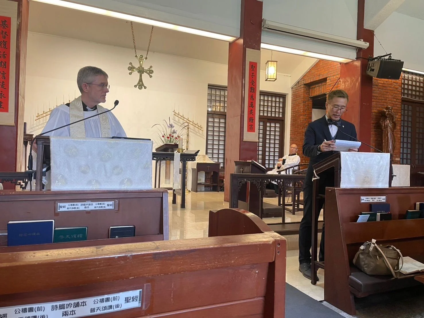 A religious ceremony inside a church with a priest and a man in a suit standing at the lecterns. The priest is dressed in white robes with a stole, and the man in a tuxedo is reading from a paper. There are decorative elements including a cross, bann