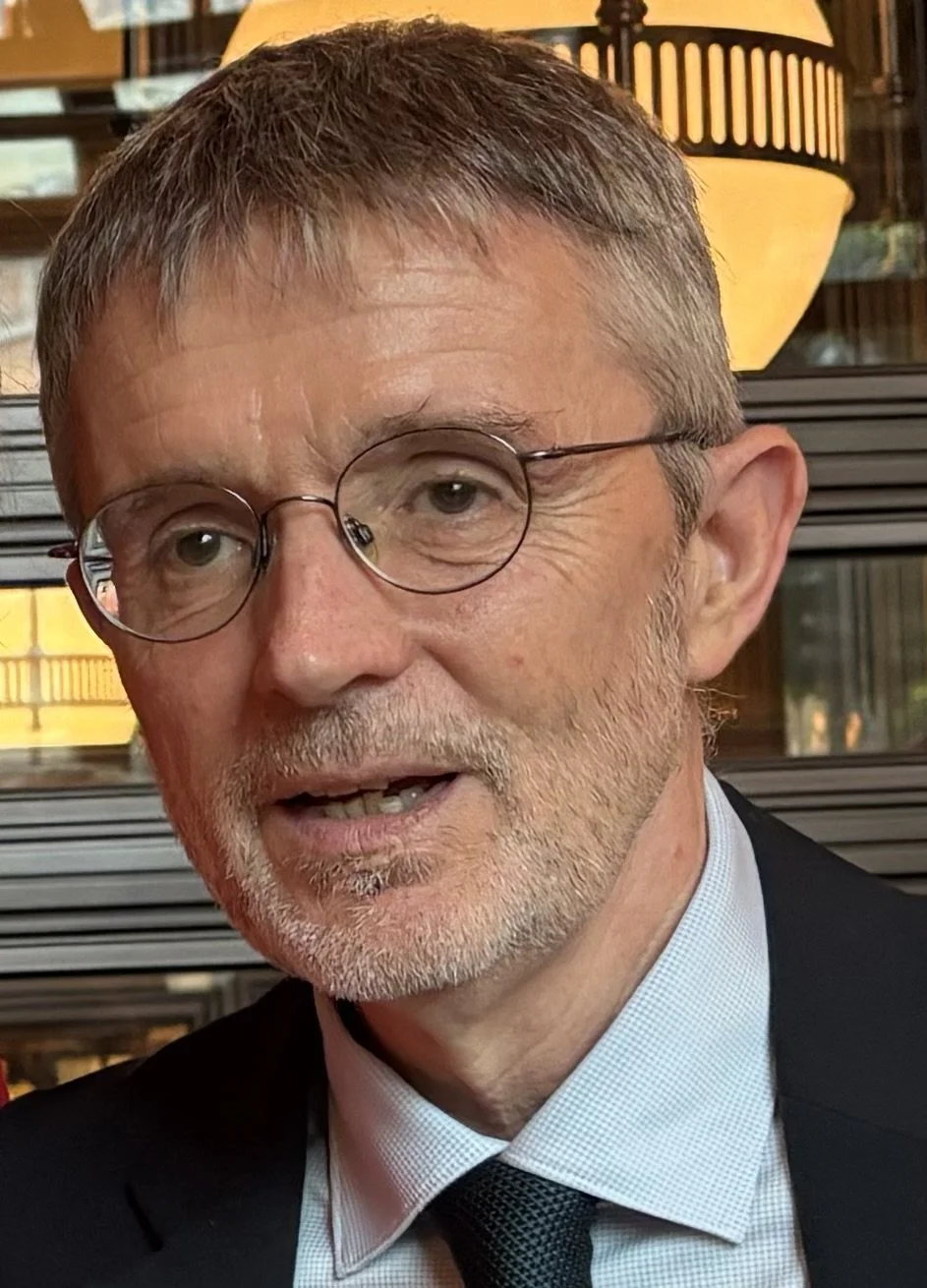 Close-up of a middle-aged man with glasses, gray hair, and a beard, wearing a suit and tie, in an indoor setting with warm lighting and wooden decor.