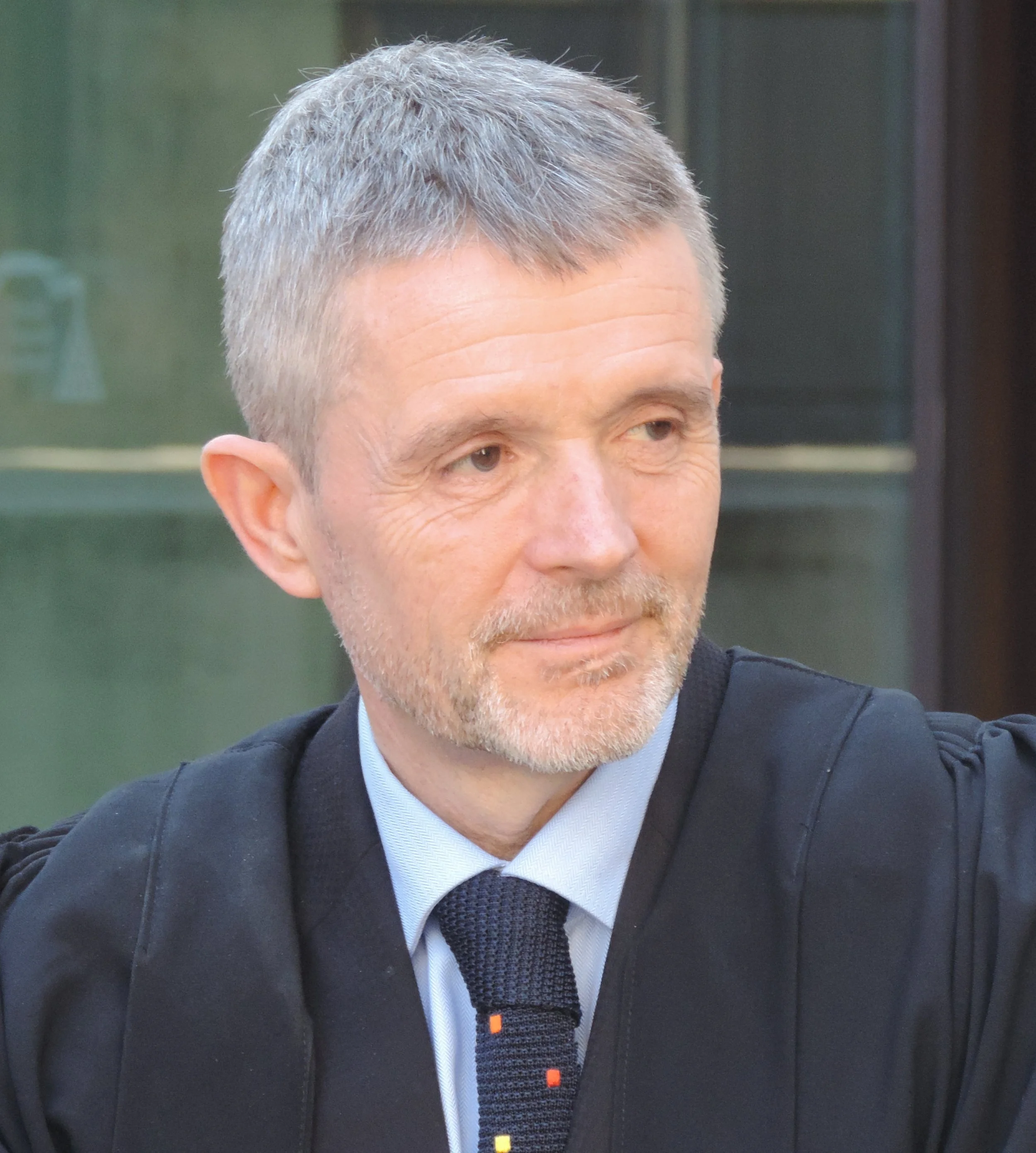Close-up of a middle-aged man with short gray hair, wearing a dark suit, white shirt, and dark patterned tie, seated outdoors with a blurred background.