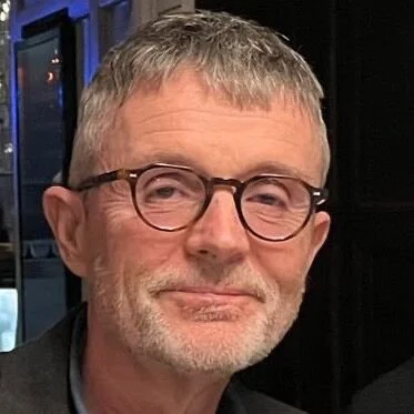 Close-up of a middle-aged man with gray hair, glasses, and a light beard smiling slightly in an indoor setting.
