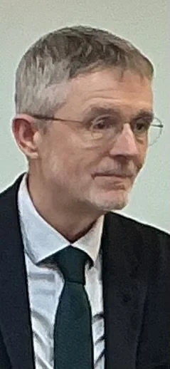 A middle-aged man with short gray hair and glasses, dressed in a black suit, white shirt, and a dark green tie, looking to the side.