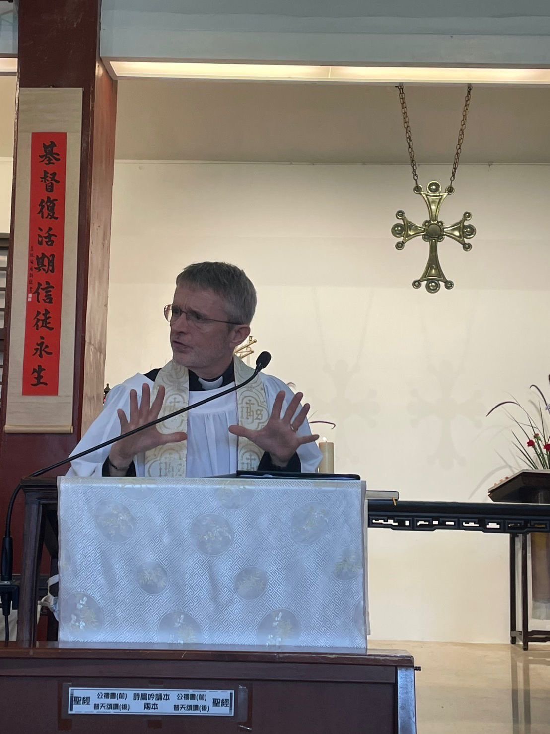 A clergyman dressed in religious robes standing behind a pulpit, speaking with hands raised, in a church setting with a cross hanging on the wall and a red vertical banner with Chinese characters.