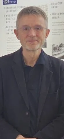A middle-aged man with glasses, short gray hair, and a trimmed beard, wearing a black suit and shirt, standing in front of a poster or informational display with text and images.