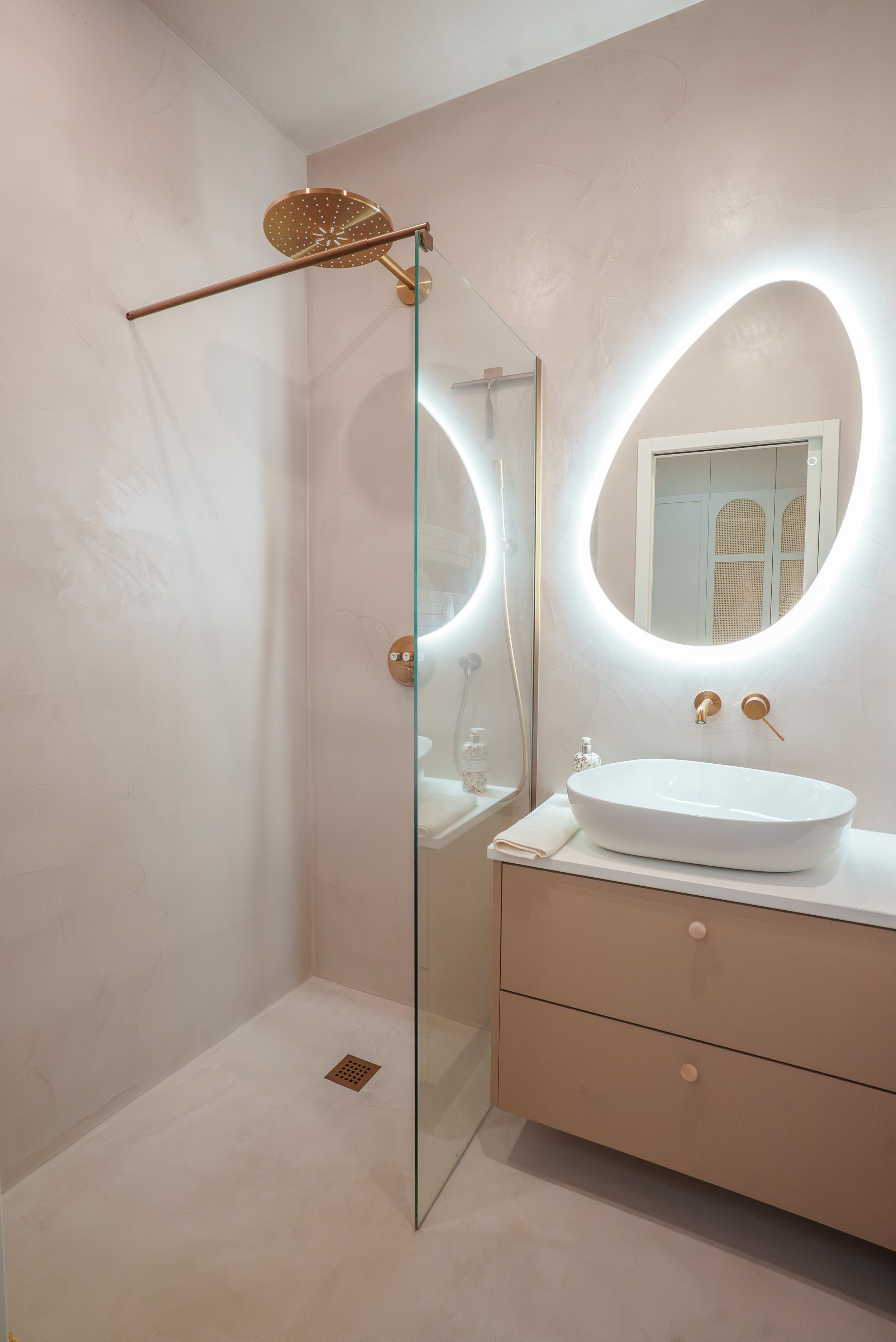 Salle de bain moderne avec un mur en verre, une douche à l'italienne, un miroir rond avec éclairage LED, un lavabo de forme ovale, un meuble beige avec tiroirs, une porte miroir et des accessoires de salle de bain en rose