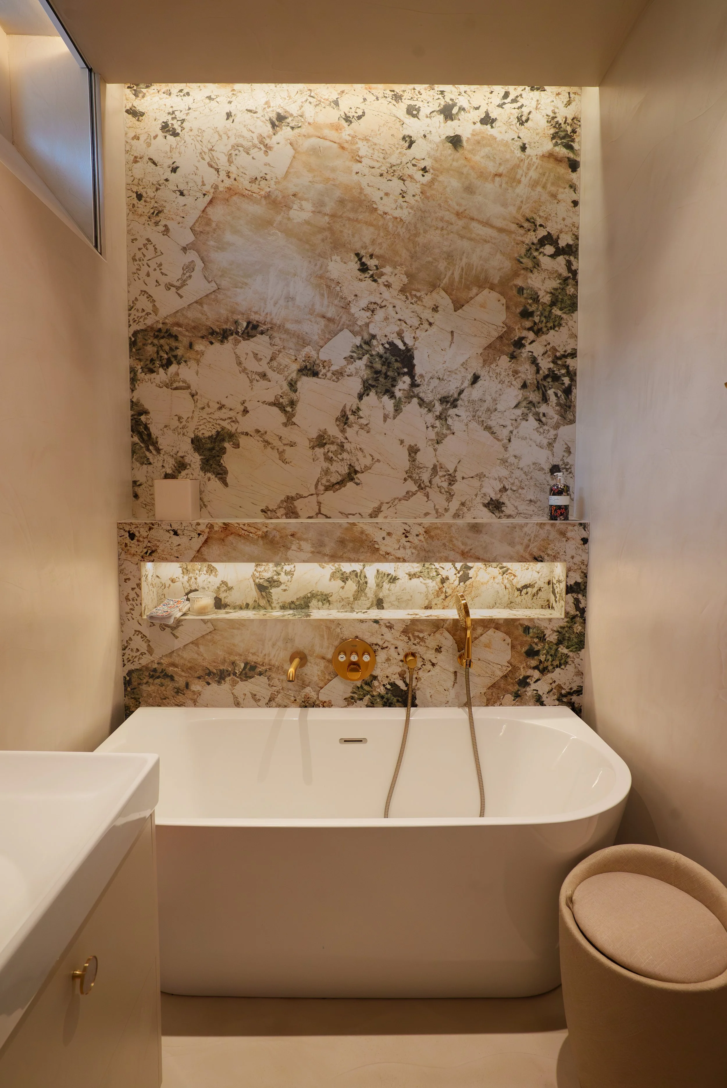 Baignoire dans une salle de bain avec mur en marbre beige et noir, avec un miroir et une petite étagère, une chaise beige en tissu et un robinet doré.
