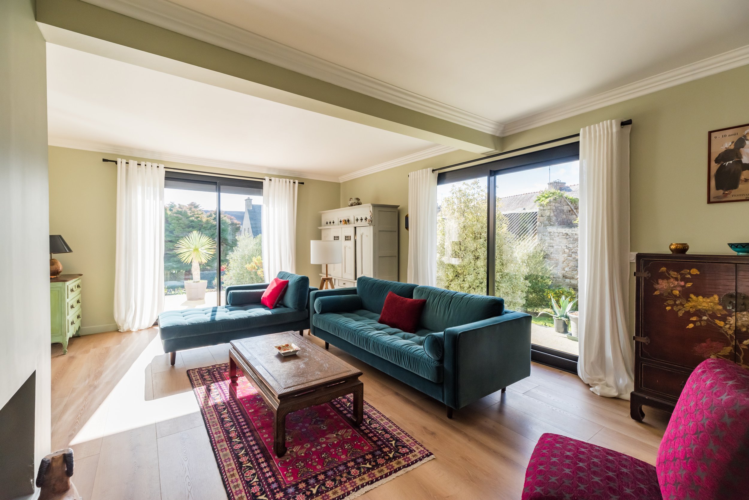 Salon lumineux avec deux canapés bleus, une table en bois, un tapis coloré, des fenêtres avec rideaux blancs laissant entrer la lumière naturelle, décorations variées et vue sur un jardin extérieur avec des arbres et un mur en pierre.