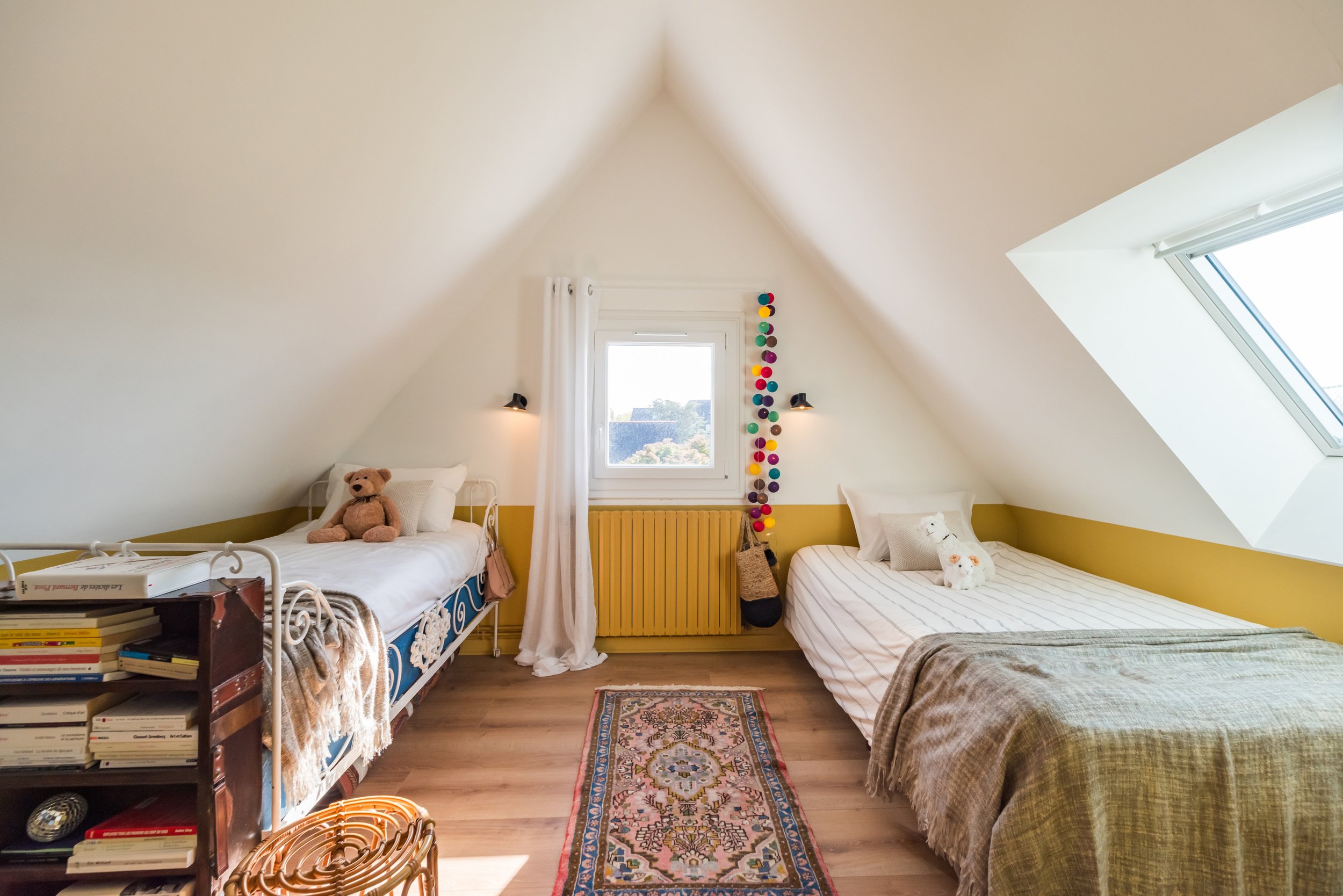 Chambre avec deux lits simples, un au gauche et un à droite, décorée avec des peluches, une bibliothèque avec des livres, un tapis coloré au sol, un mur jaune, une fenêtre avec jour, un mur blanc, un plafond en pente avec une lucarne, et des lumières