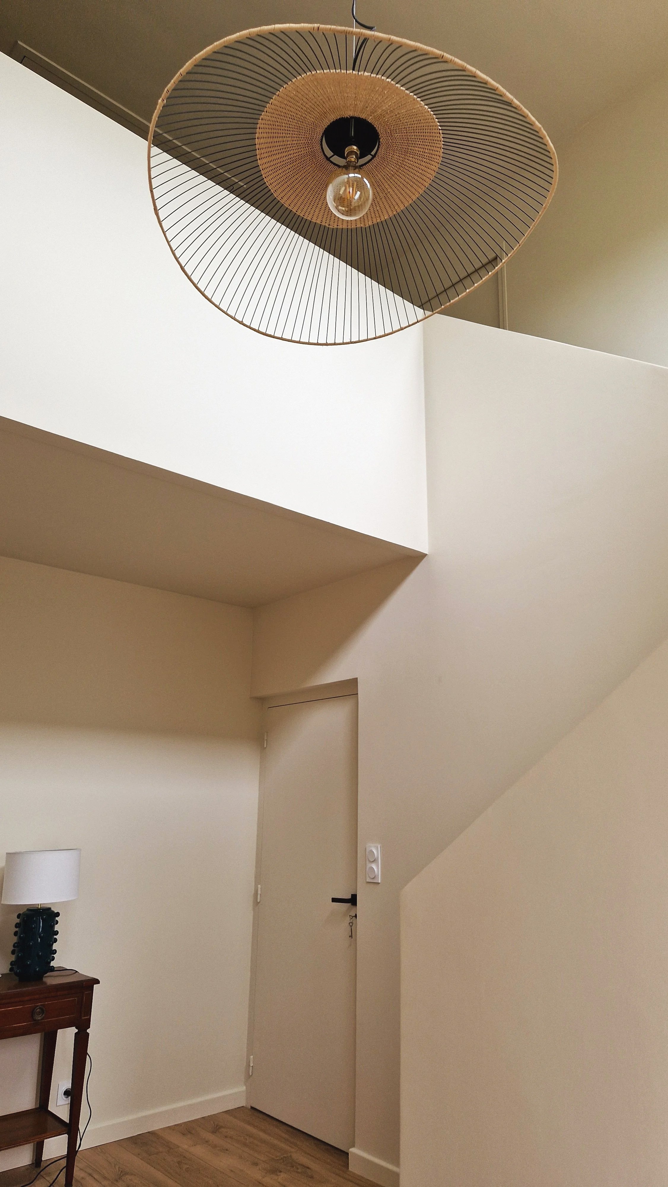 Plafonnier design suspendu au plafond, éclairage dans une pièce intérieure avec mur blanc et coin salon, porte et interrupteur visible.