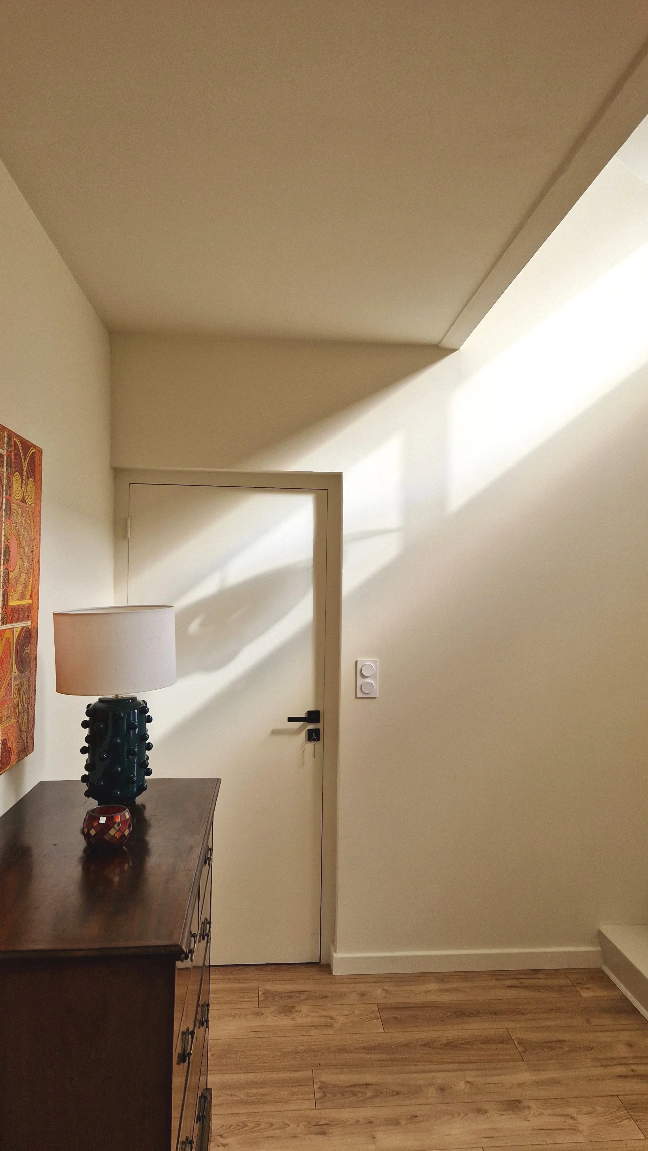 Une entrée dans une maison avec une porte blanche, une lampe sur une table, un tableau coloré sur le mur gauche, et un sol en bois,