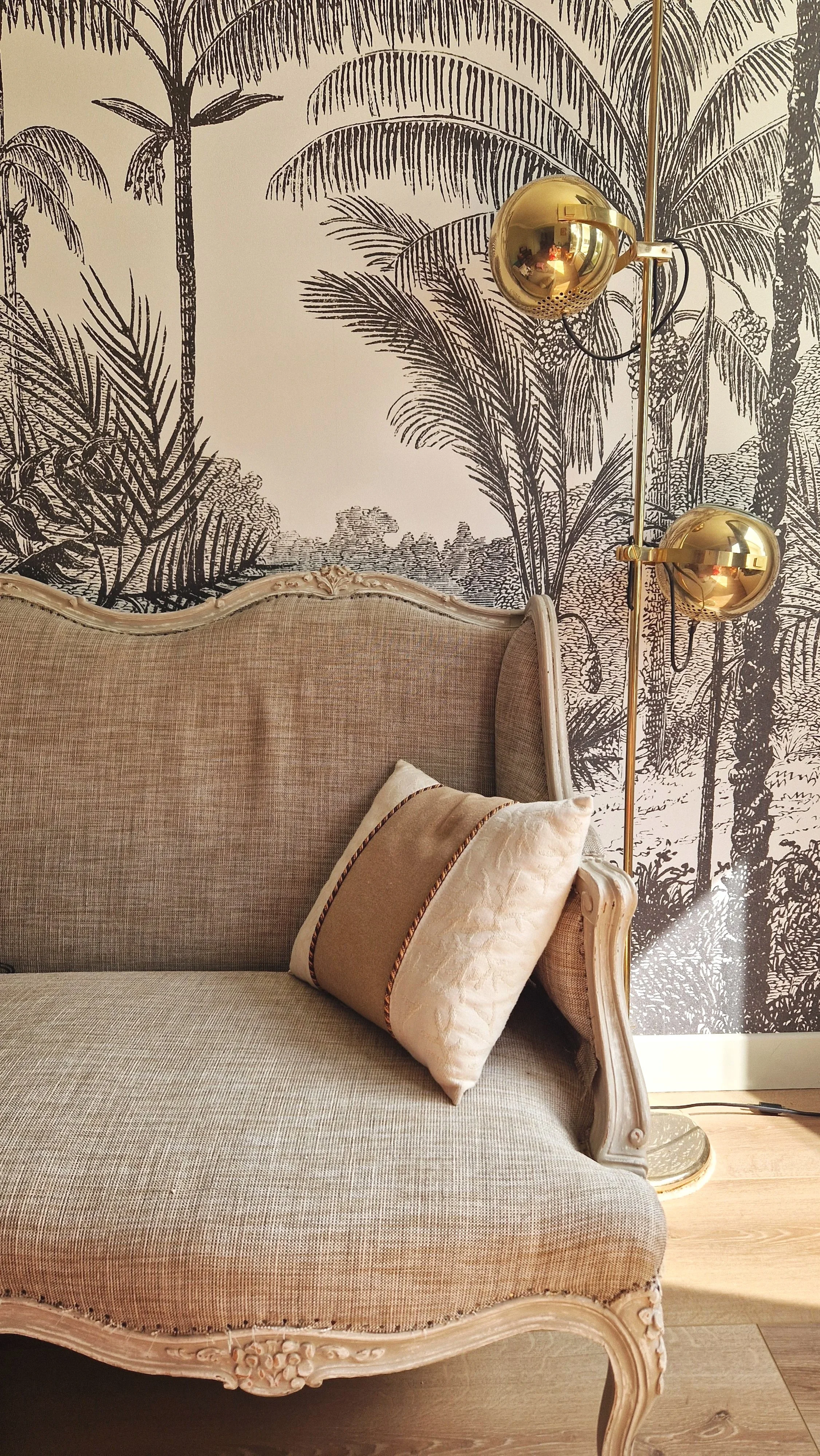 Un canapé beige avec une housse en tissu, un coussin crème avec bordure en fil métallique, un mur avec un papier peint à motif de palmiers noir et blanc, et une lampe en laiton avec deux sphères dorées.