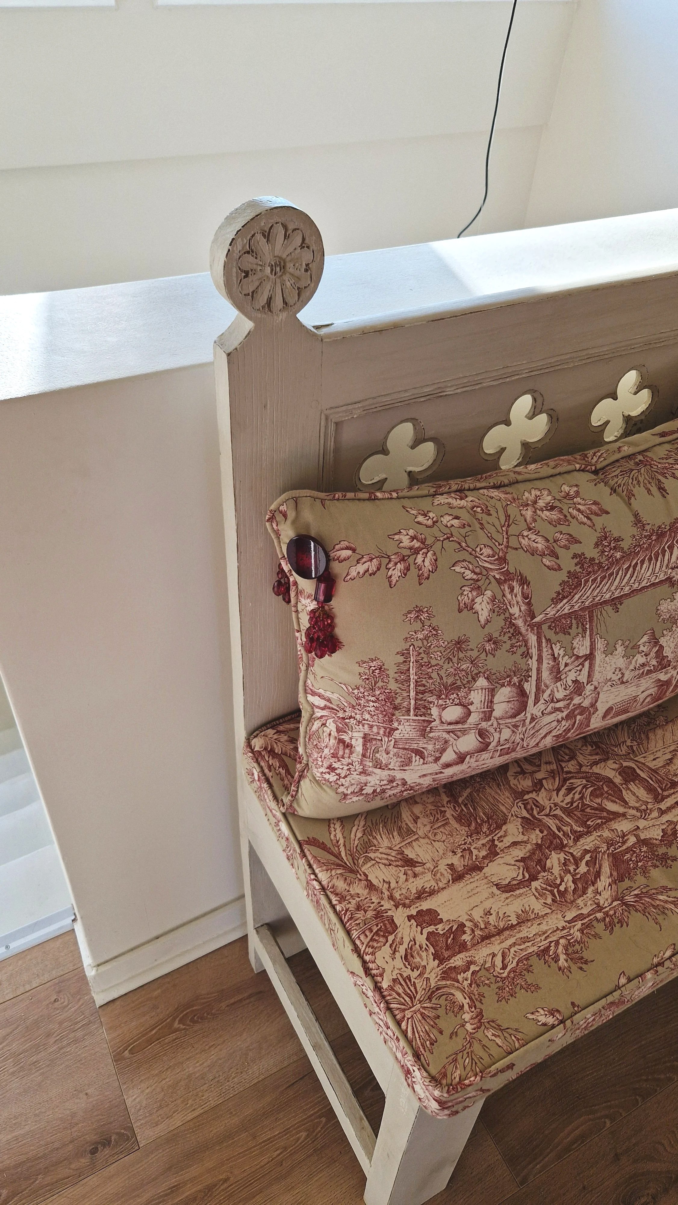 Un lit en bois blanc avec une tête de lit ornée de découpes en forme de trèfle et une housse de coussin beige avec un motif baroque rouge, posé sur un sol en bois.