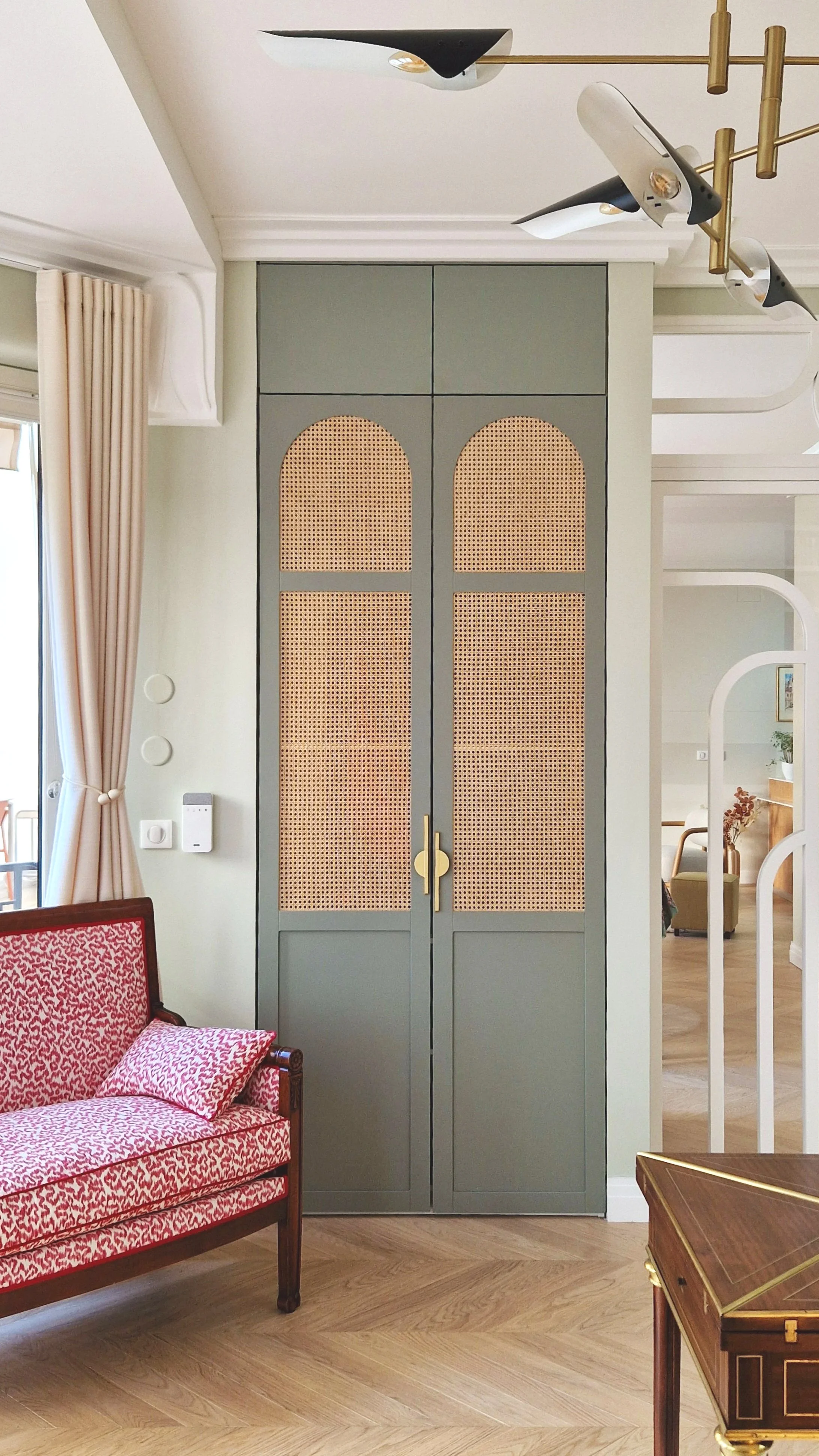 Un mur dans un salon avec une armoire grise équipée de portes en cannage. À gauche, un canapé en bois avec coussins à motif rouge et blanc, à droite une table en bois avec détails en or. On distingue également une baie vitrée avec rideaux crème et un
