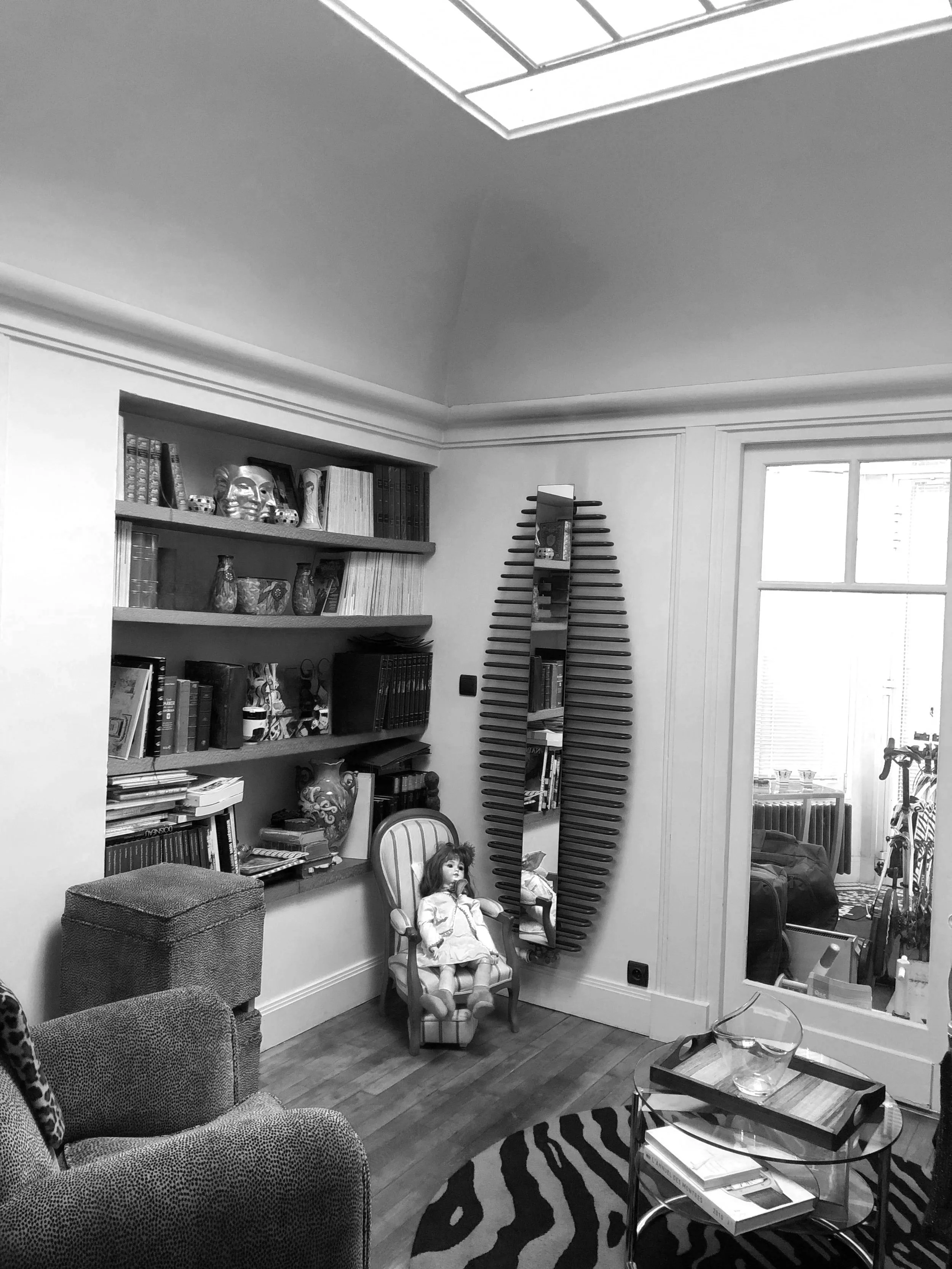 Salon avec étagère remplie de livres, décoration et une poupée dans un fauteuil, vue d'une fenêtre et d'une porte vers un balcon ou une pièce adjacente.