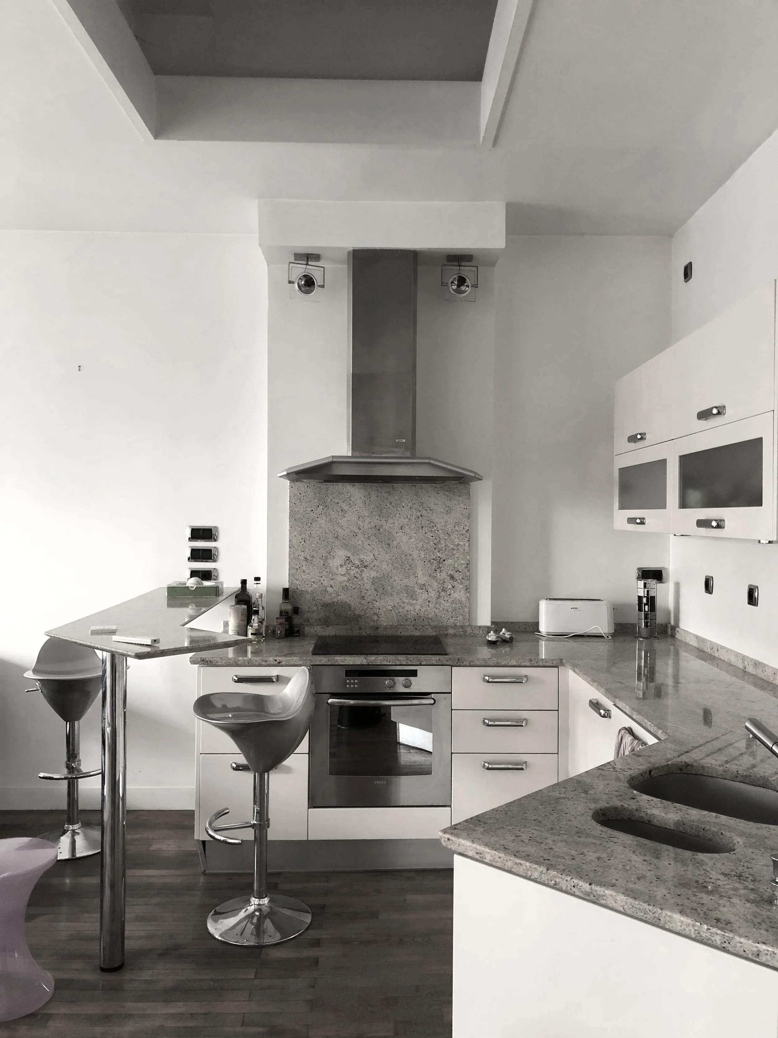 Une cuisine moderne avec comptoir en granite, armoires blanches et électroménagers en acier inoxydable. Il y a un bar avec deux tabourets modernes, une hotte aspirante au-dessus de la cuisinière, et diverses bouteilles et objets sur le contreplaqué d