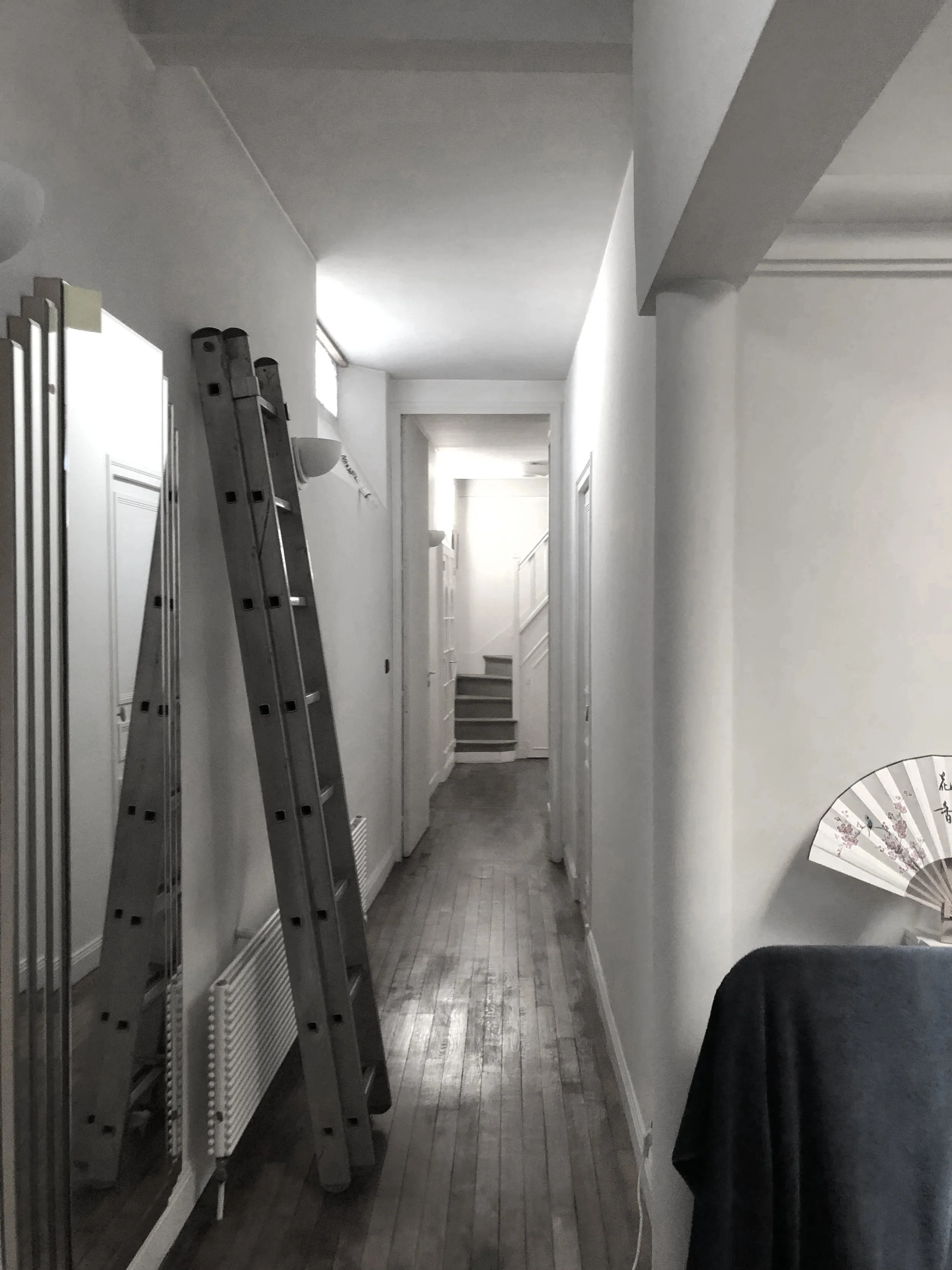 Couloir intérieur d'une maison avec un escalier en arrière-plan, deux échelles en porte-à-faux sur le côté gauche, une fenêtre en hauteur laisse entrer la lumière, un radiateur blanc au bas du mur à gauche, un éventuel ventilateur ou radiateur sur la