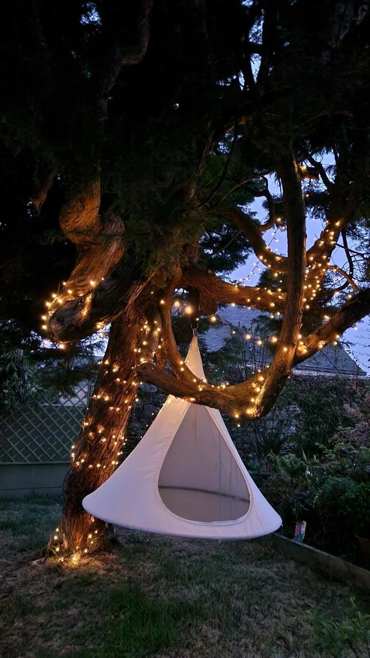 Une tente blanche suspendue entre les branches d'un arbre éclairé par des guirlandes lumineuses, dans un jardin au crépuscule.