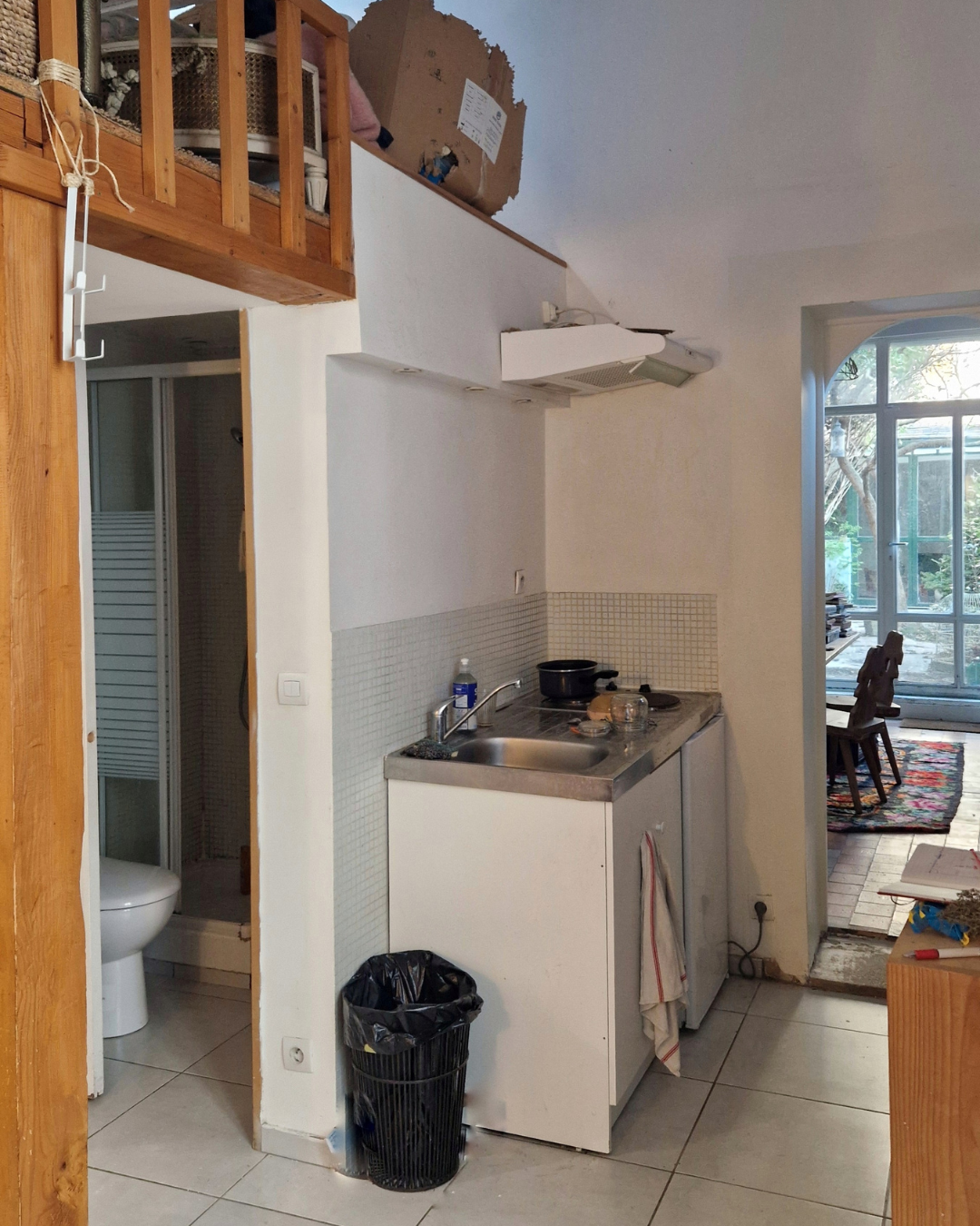 Petite cuisine avec évier, plaque de cuisson et quelques ustensiles. Cheminée en haut, porte menant à une salle de bains avec WC, et vue vers une pièce avec des chaises et une grande fenêtre