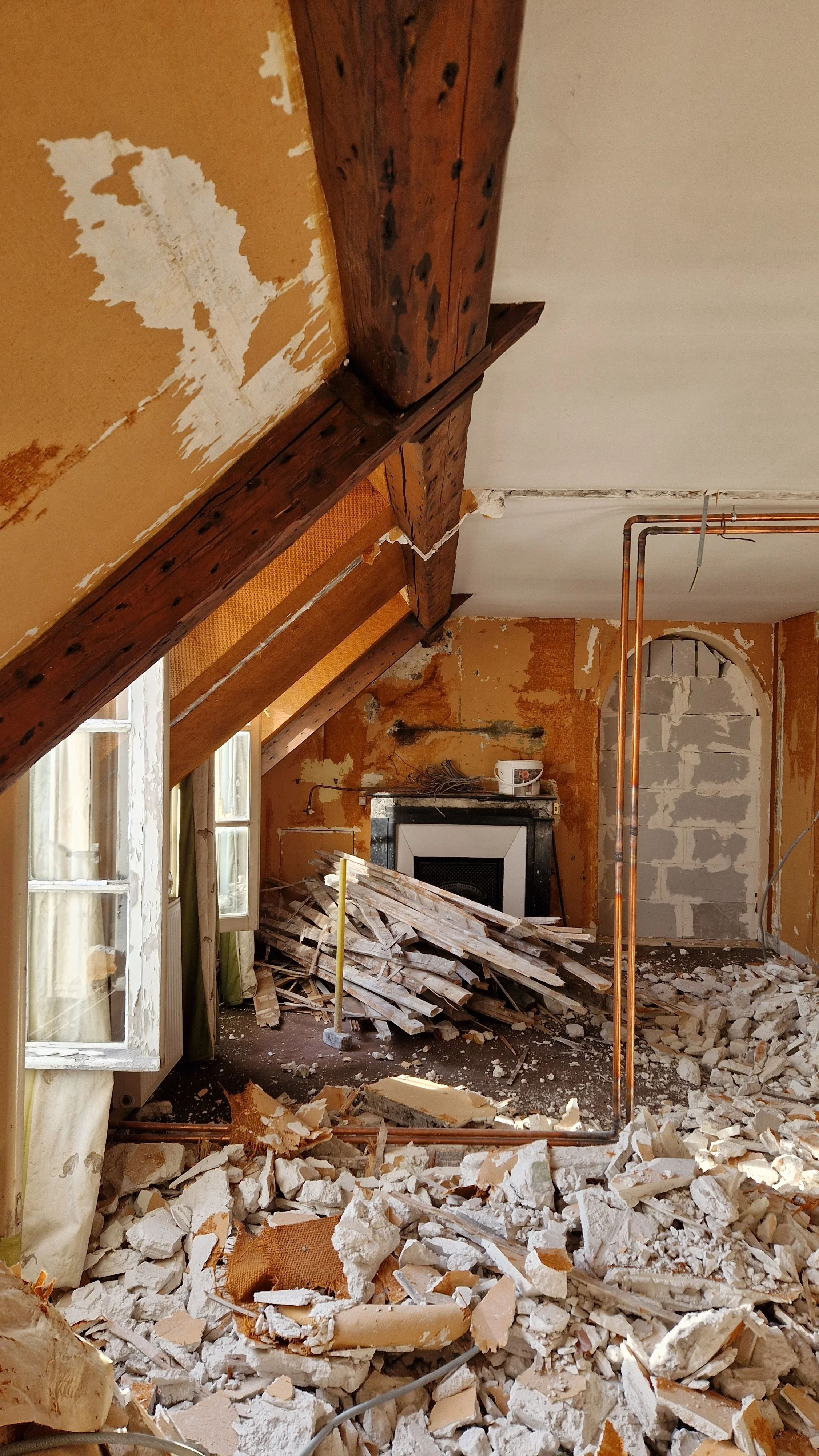 Une pièce en rénovation avec des poutres en bois au plafond, des murs partiellement déchirés, et un tas de débris de plâtre au sol, avec une cheminée en arrière-plan.